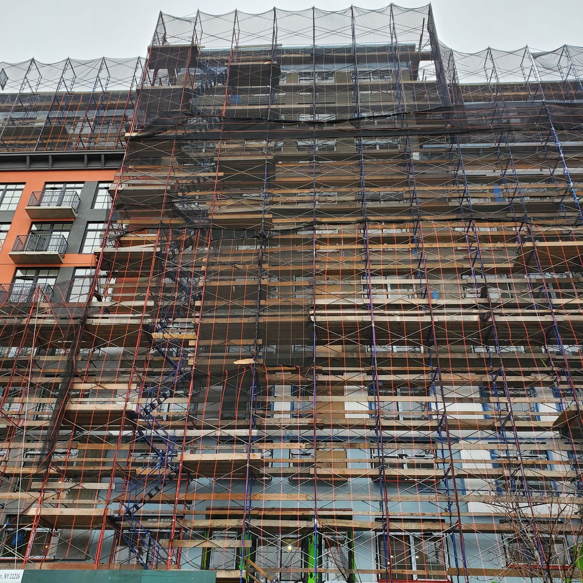 Building under construction, covered in scaffolding and safety netting; overcast sky.