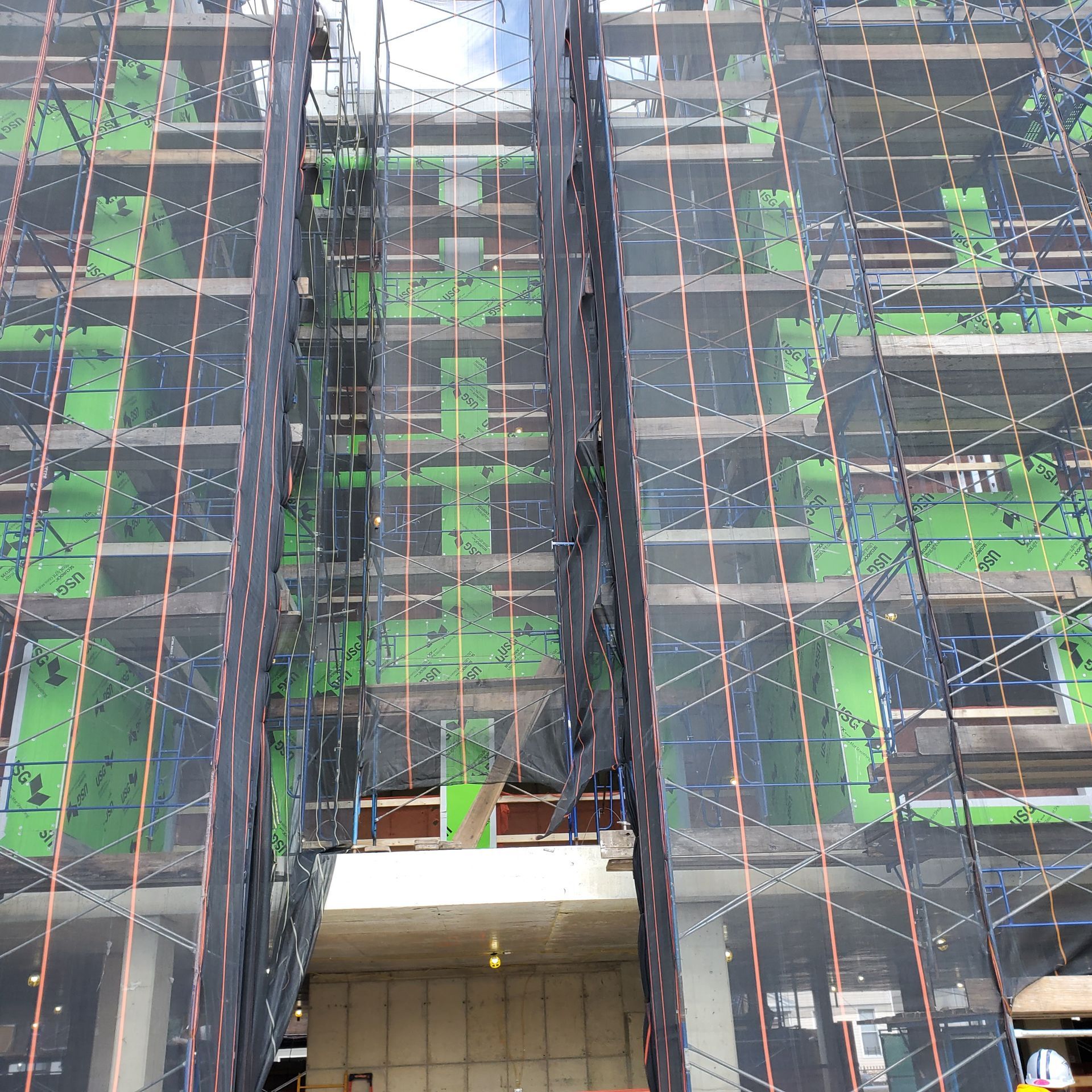 Building under construction, covered in scaffolding and black netting; green wall sections visible.