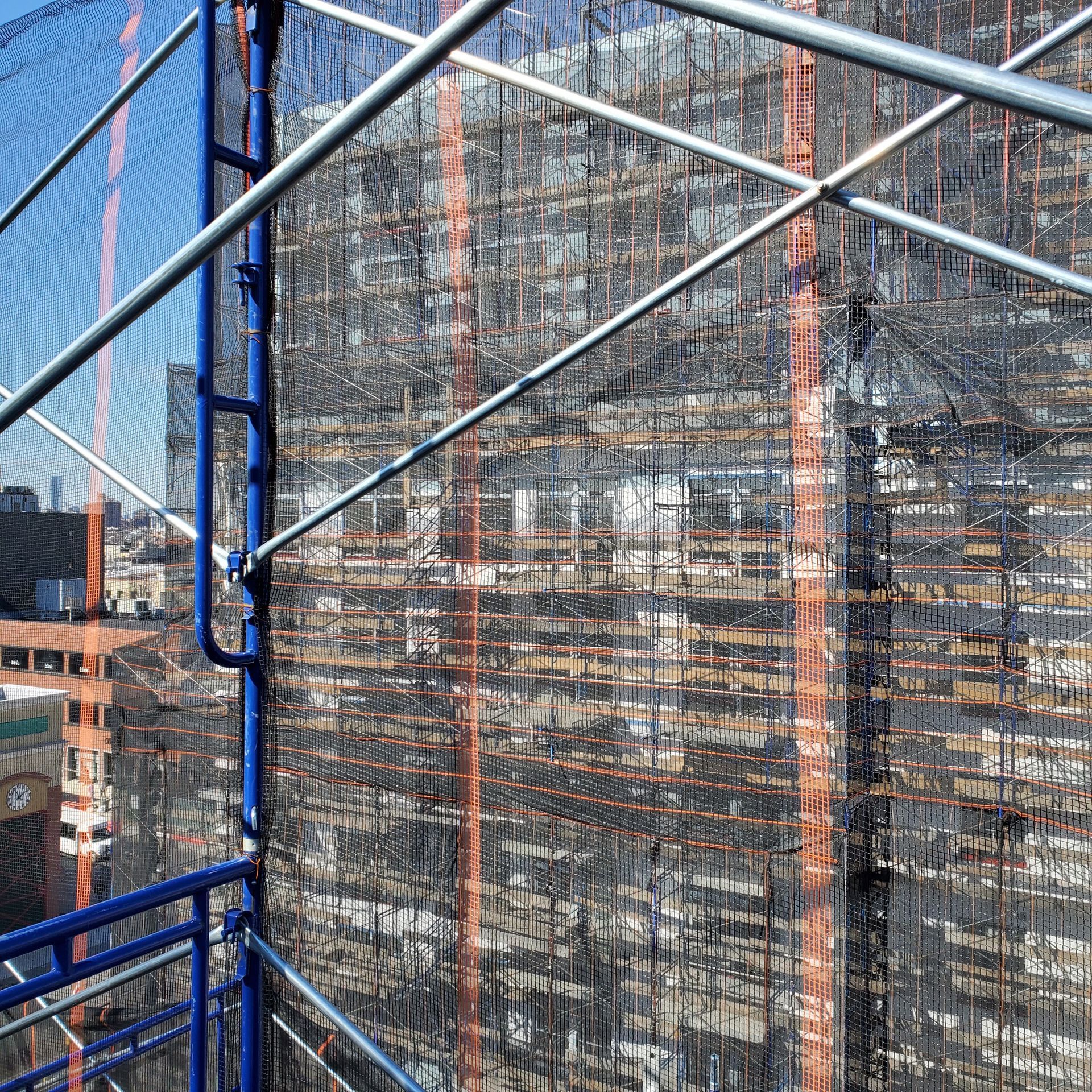 Scaffolding on a building covered in black mesh with blue and silver supports; city in the background.