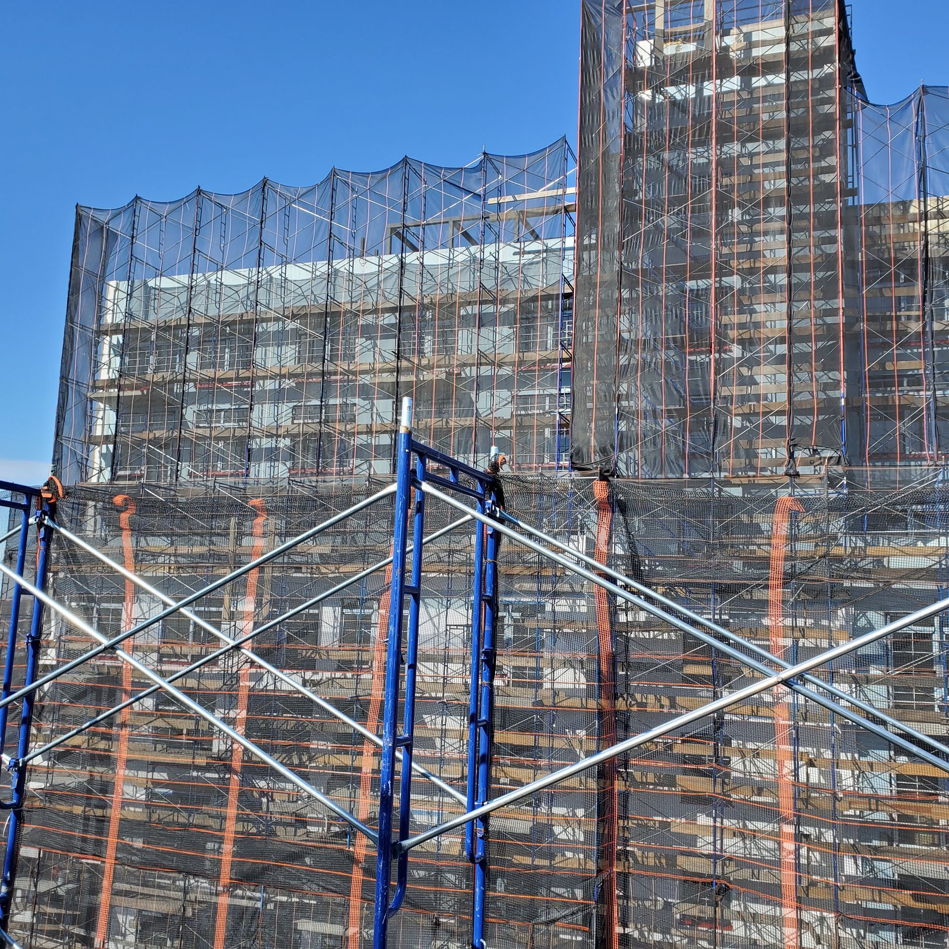 Building under construction, encased in scaffolding and protective netting against a blue sky.