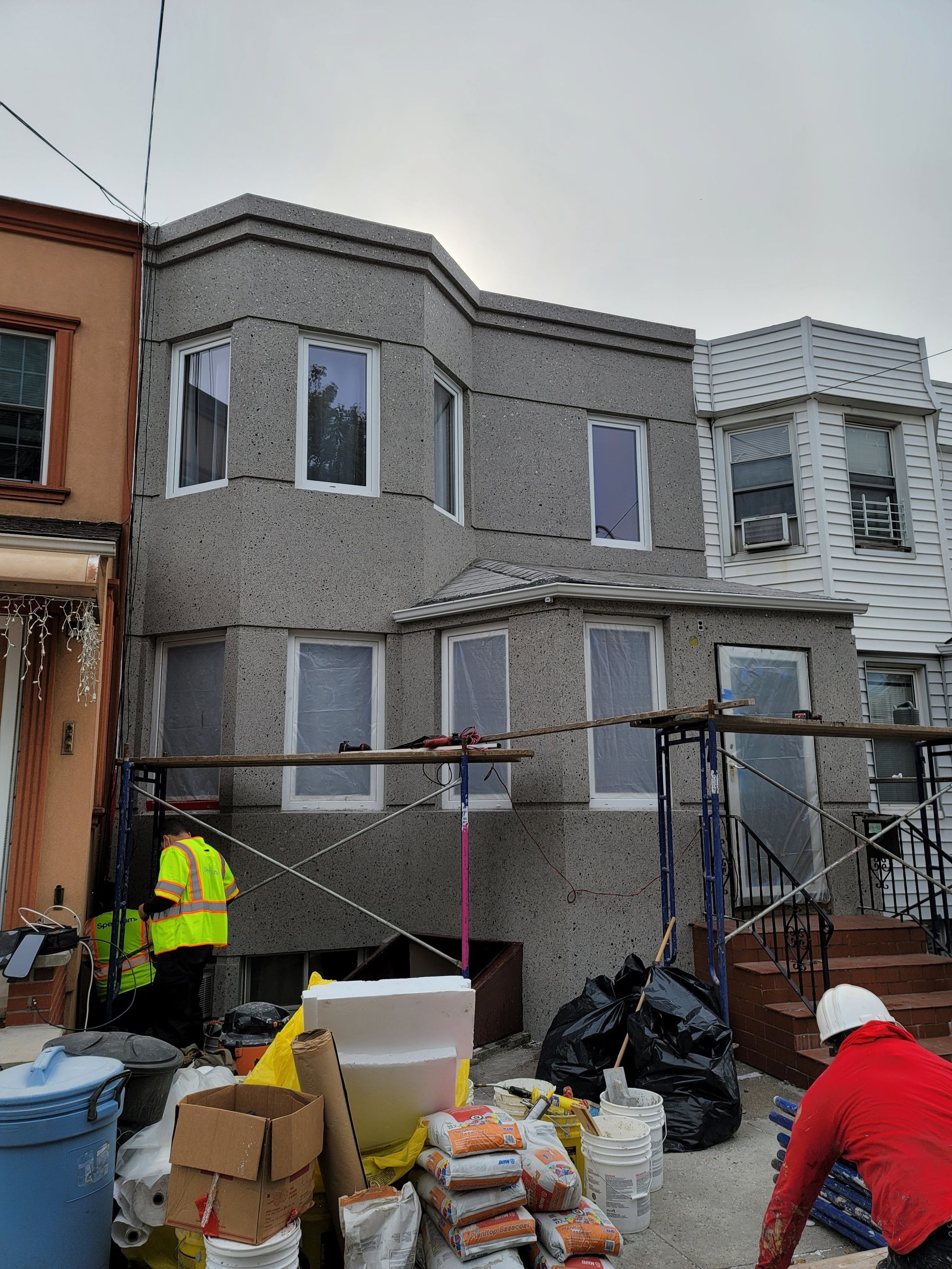 Two-story house under construction with scaffolding and workers. Gray stucco exterior with bay windows.