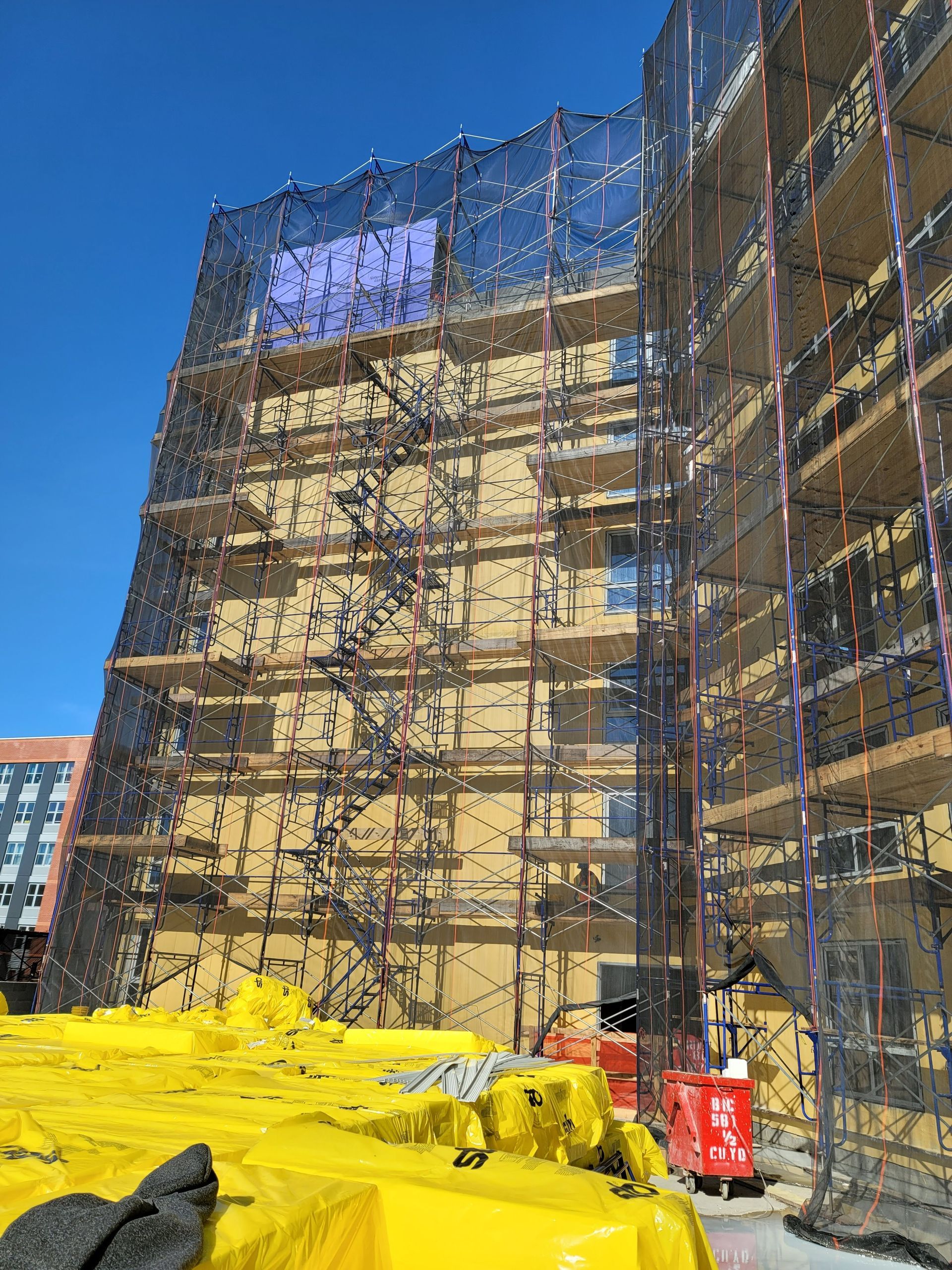 Building under construction, covered in scaffolding and safety netting, with yellow tarps below.