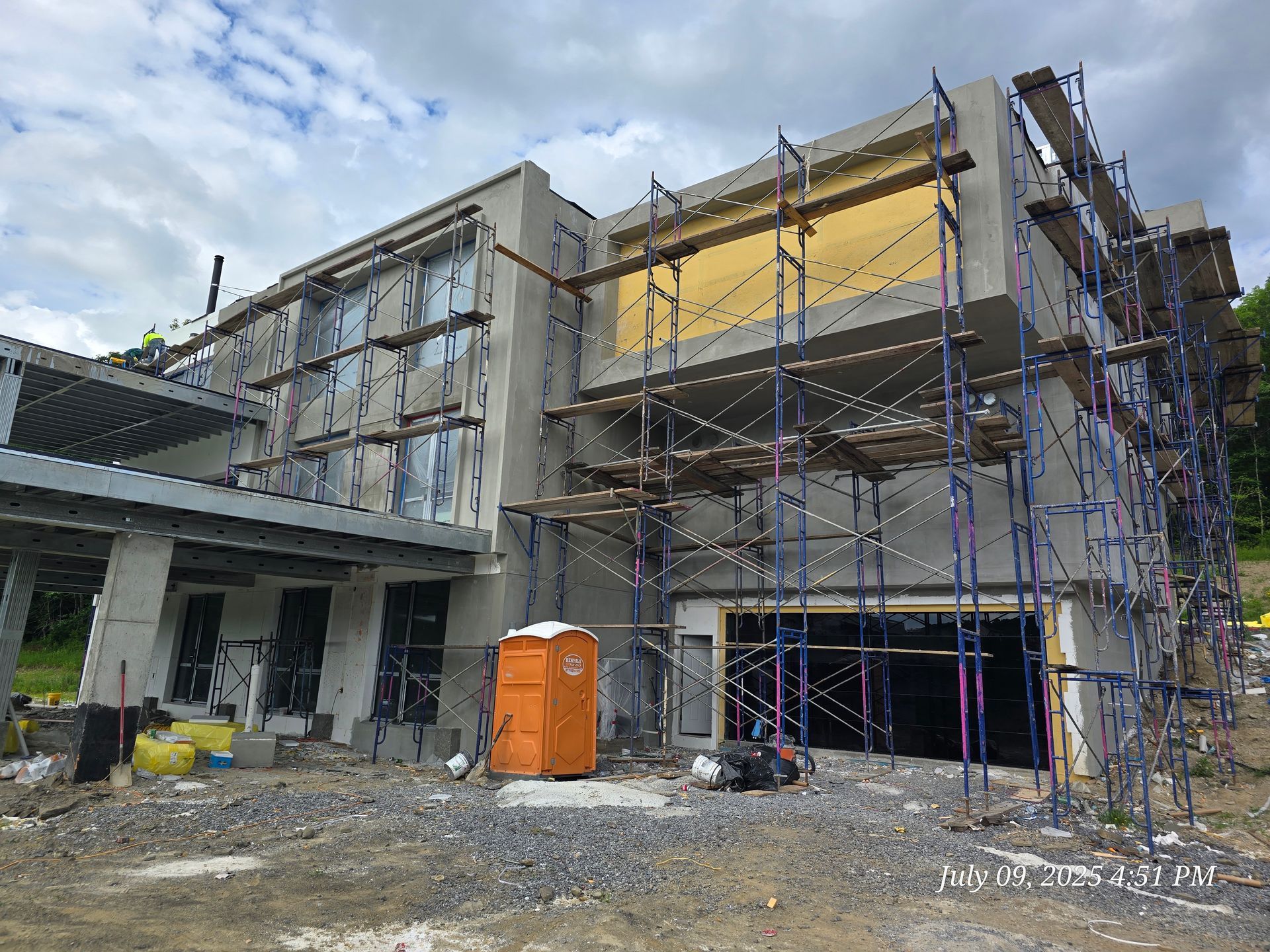 Building under construction with scaffolding and orange portable toilet. Concrete and gray exterior.