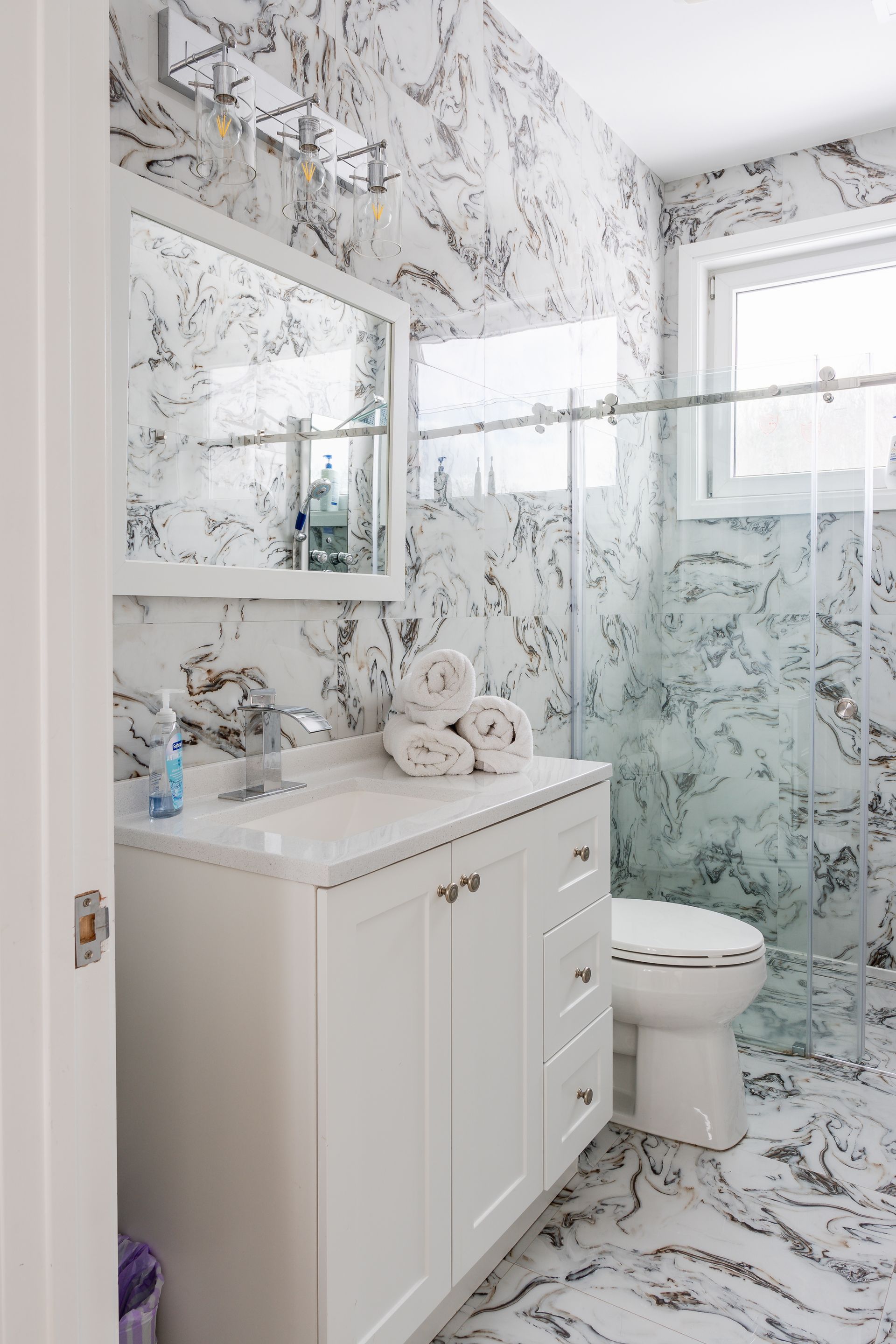 White and marble-tiled bathroom with vanity, toilet, shower, and window.