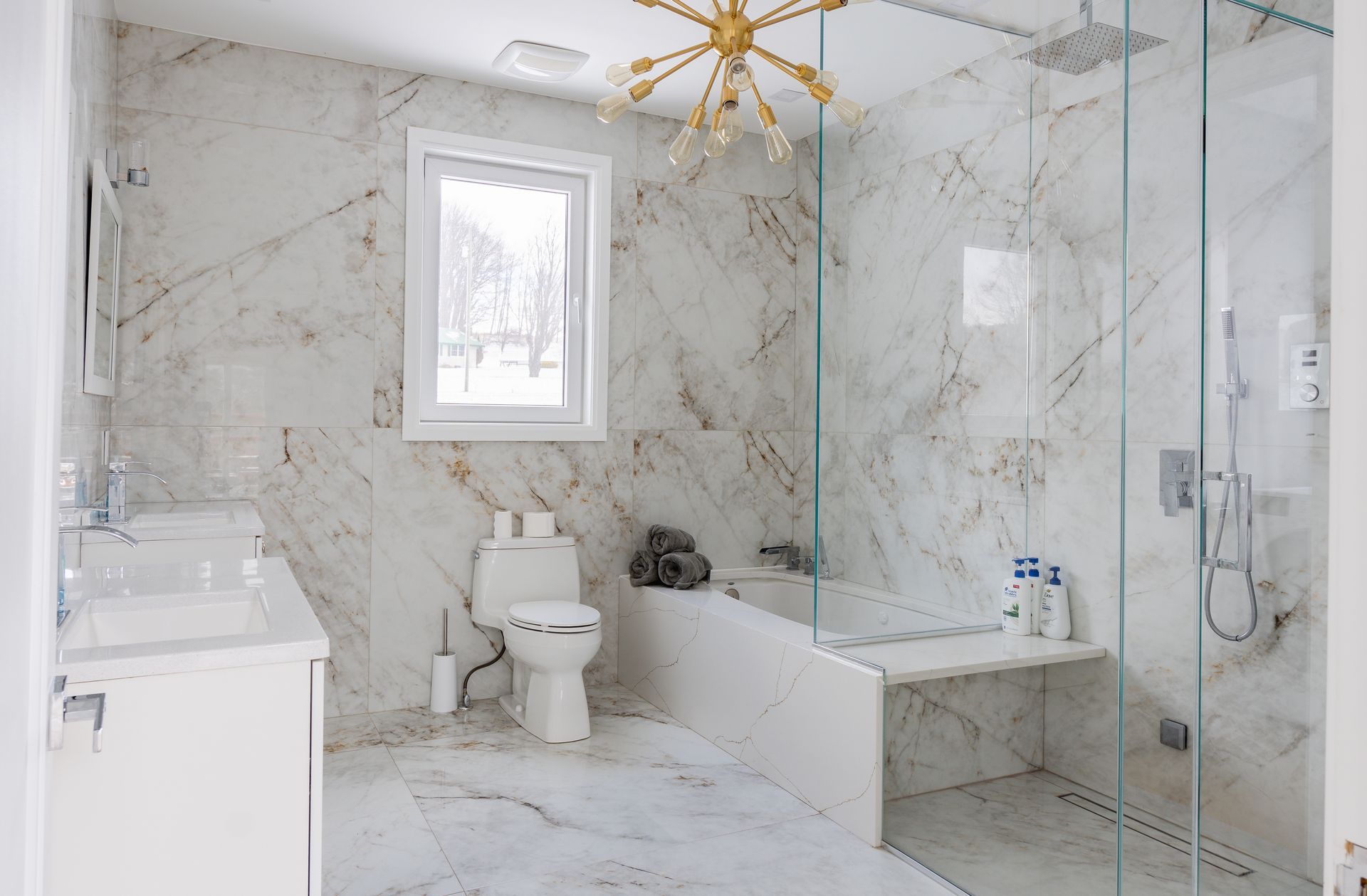 Luxurious bathroom with marble walls, glass shower, toilet, window, and gold chandelier.