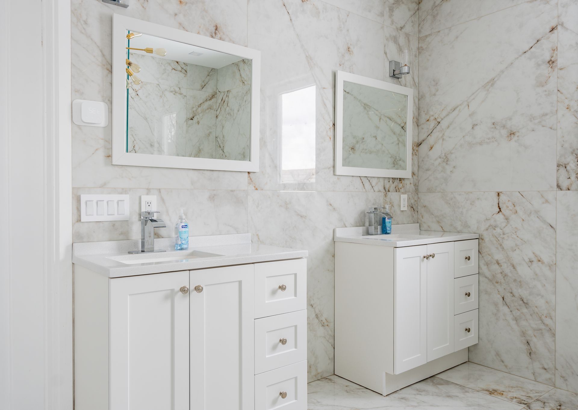 Two white vanities with mirrors, against marble tile walls, in a bathroom.