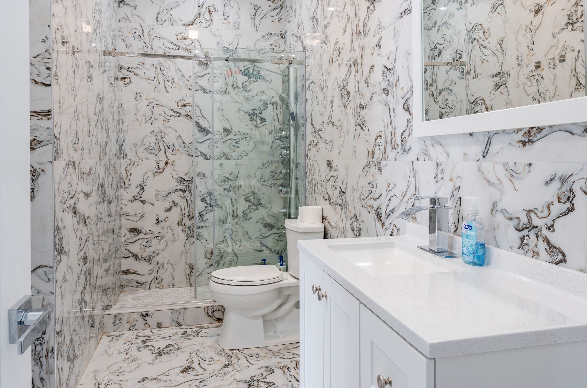 Bathroom with marble-patterned walls and floor. White vanity, toilet, and shower with glass door.
