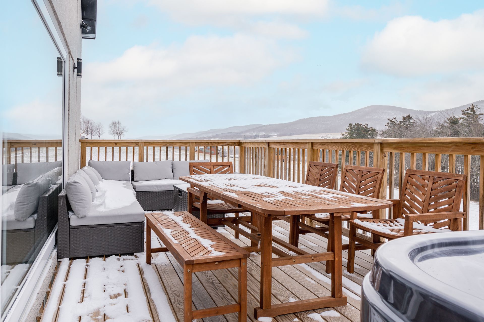 Snow-covered wooden deck with outdoor furniture: sectional sofa, dining table, chairs, and hot tub, with a mountain view.