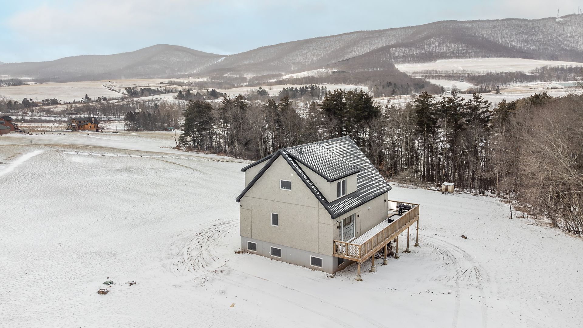 House in a snowy field with mountains in the background. A wooden deck and trees are nearby.