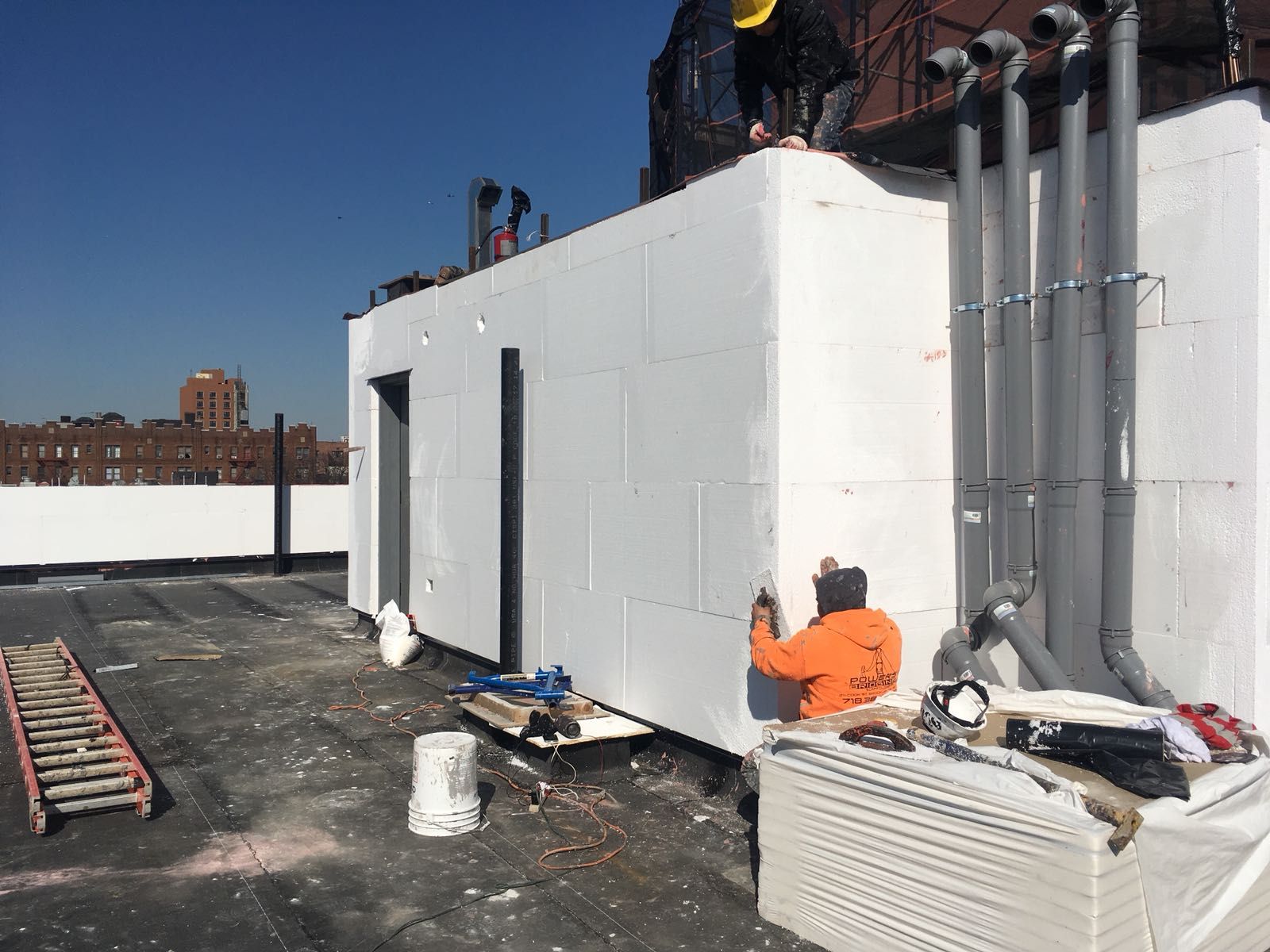Construction workers on a rooftop building white insulated walls, pipes, with tools and materials. Blue sky.