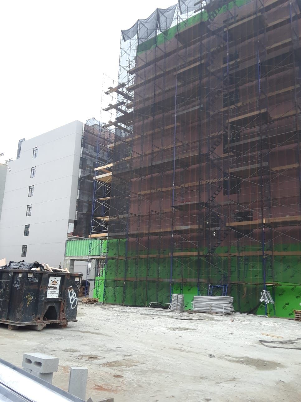 Construction site with scaffolding and netting on a multi-story building. Dumpsters and debris in the foreground.