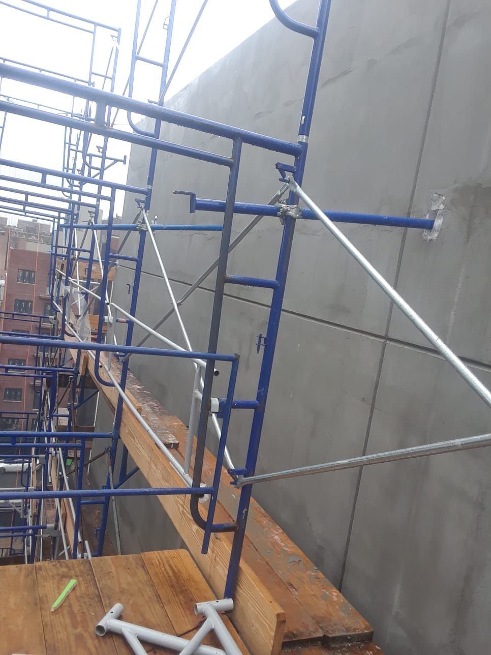 Blue scaffolding attached to a gray concrete wall, wooden plank platform.