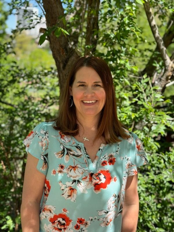A woman in a floral shirt is standing in front of a tree.