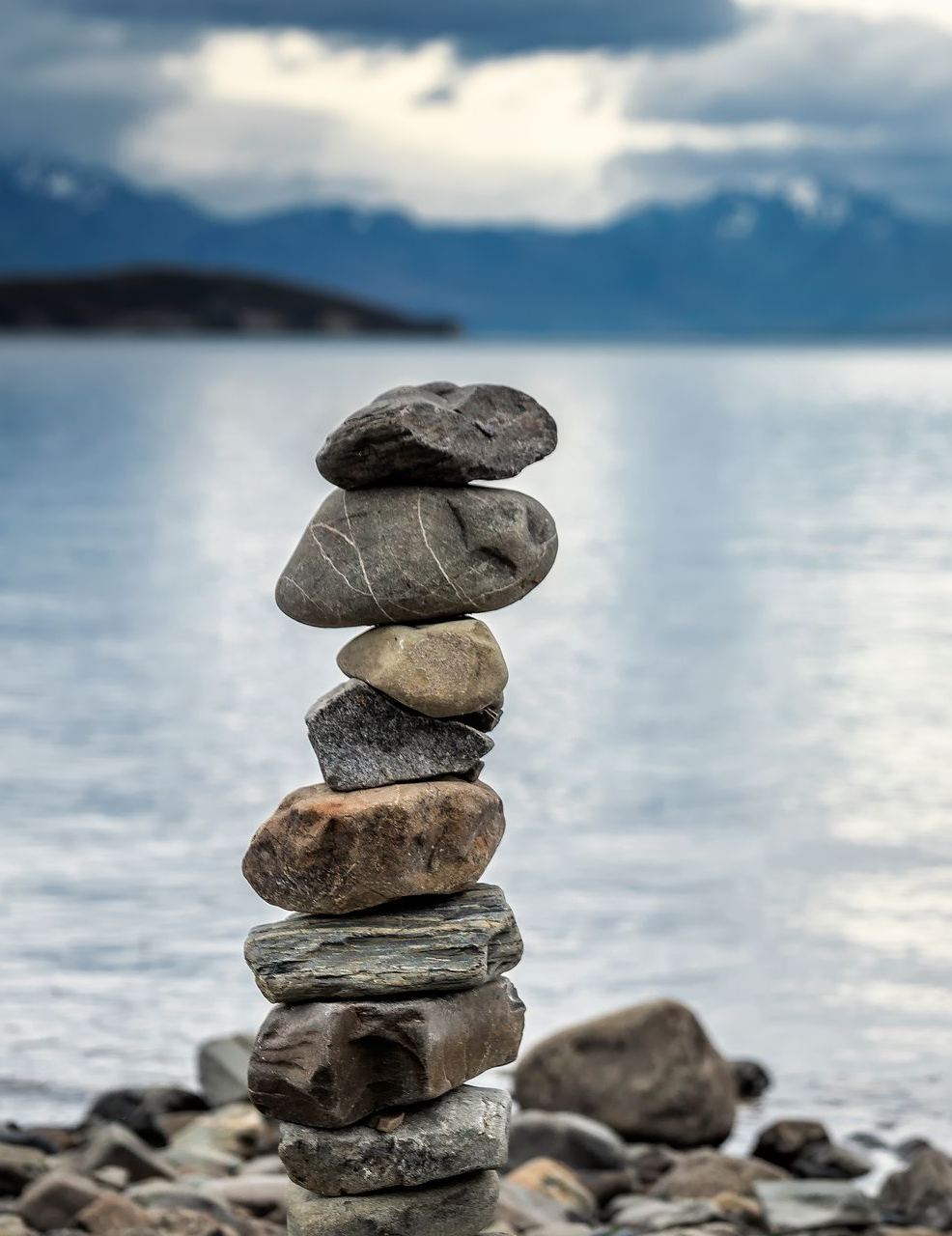 Stacked stones on a rocky shore beside a calm lake with mountains in the misty background