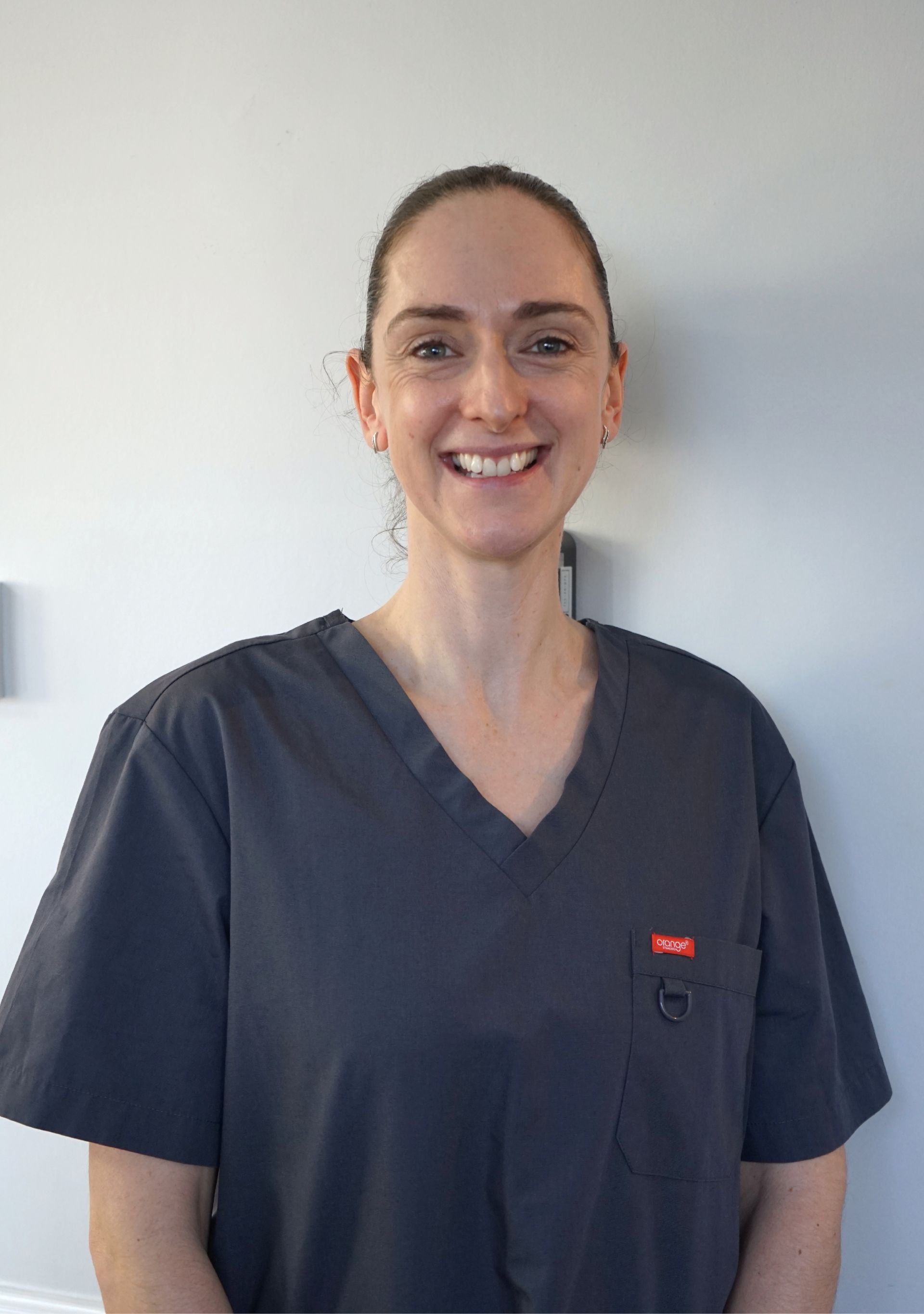 Woman in grey scrubs smiles, standing in front of a light-colored wall.