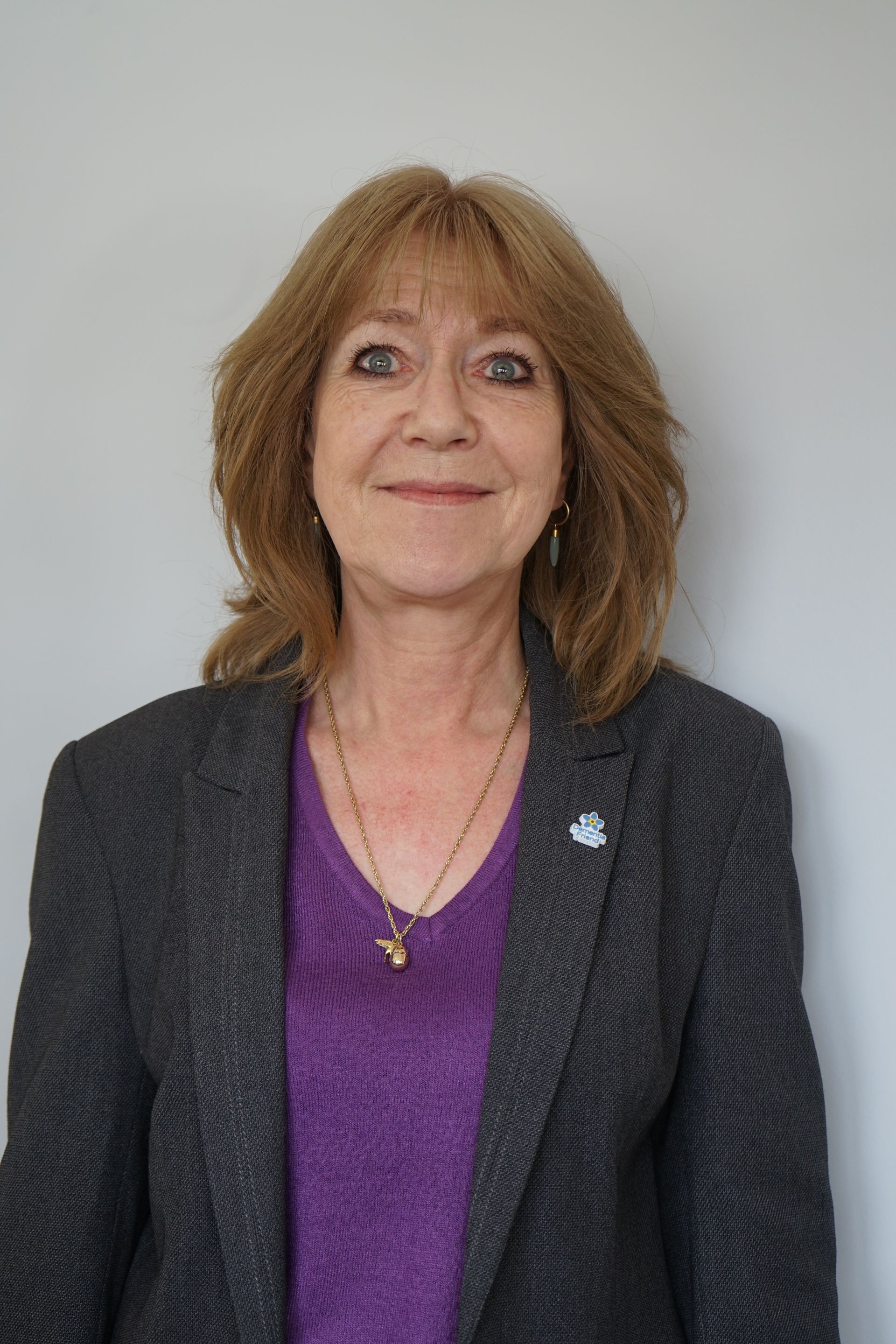 Woman with shoulder-length auburn hair, wearing a purple shirt and gray blazer, smiling, standing in front of a gray wall.