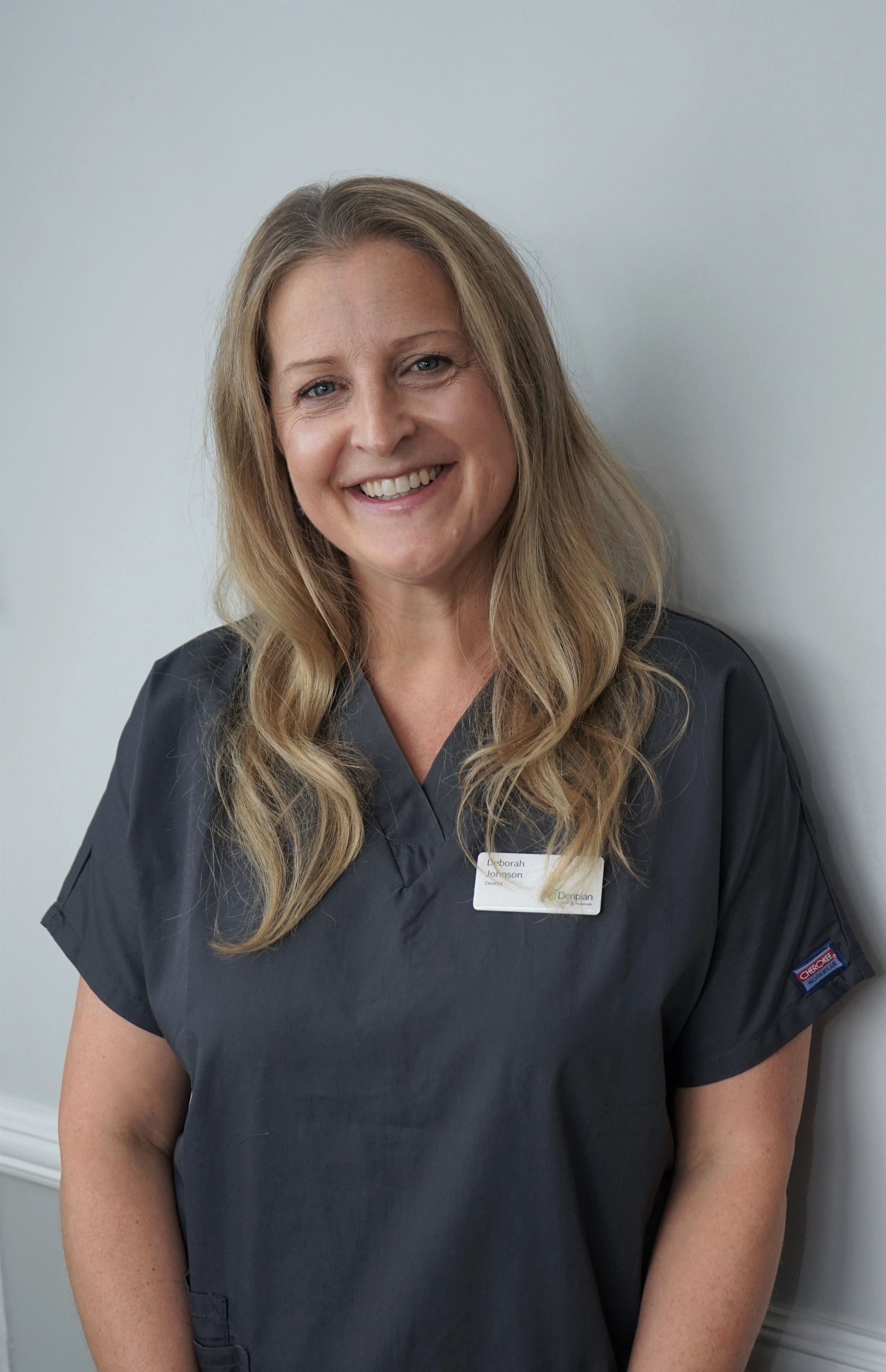 Woman in gray scrubs smiles at the camera; leaning against a light gray wall.