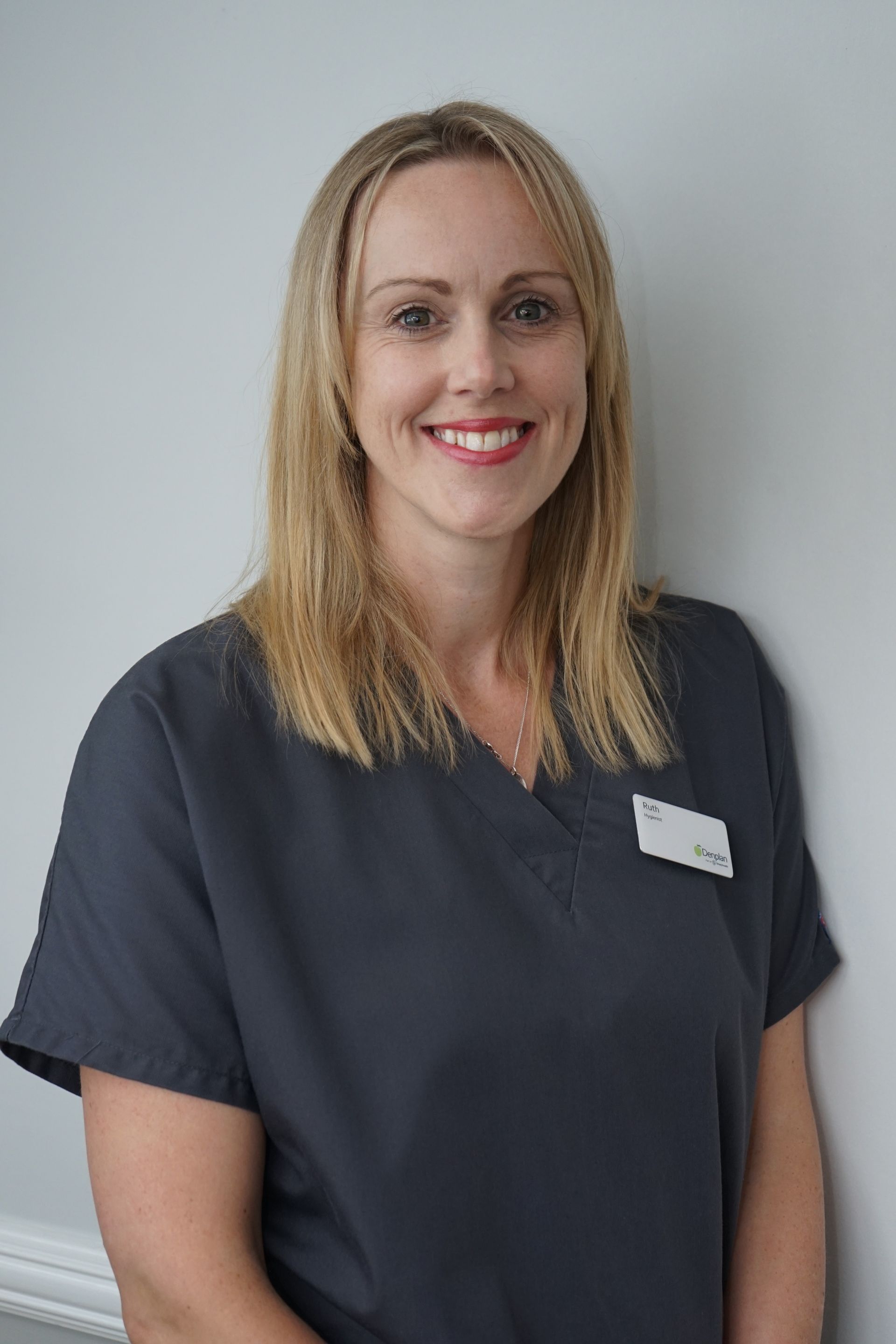 Woman with blonde hair wearing blue scrubs, smiling, leaning against a white wall.