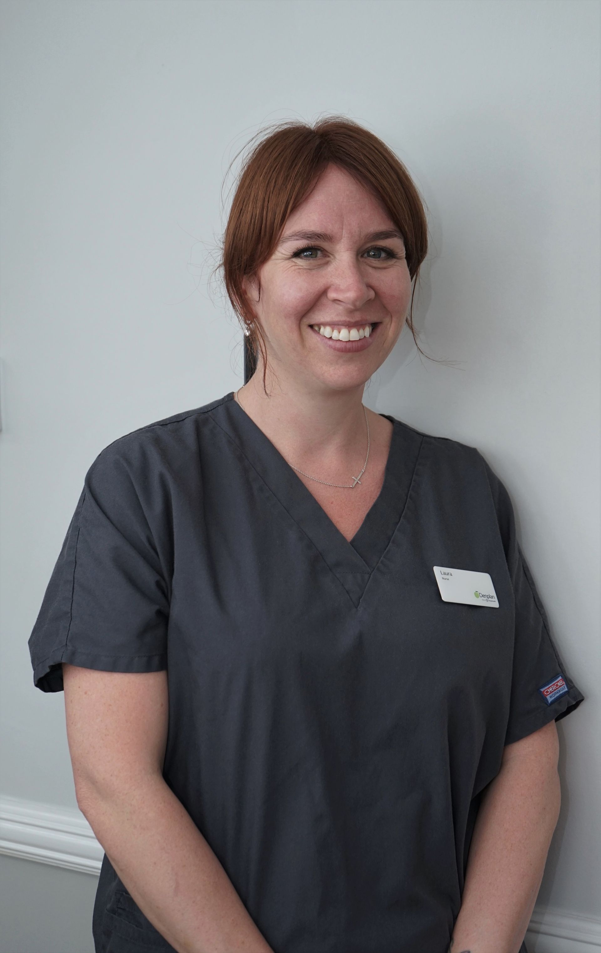 Woman with red hair smiles, wearing gray scrubs and a name tag, standing against a white wall.