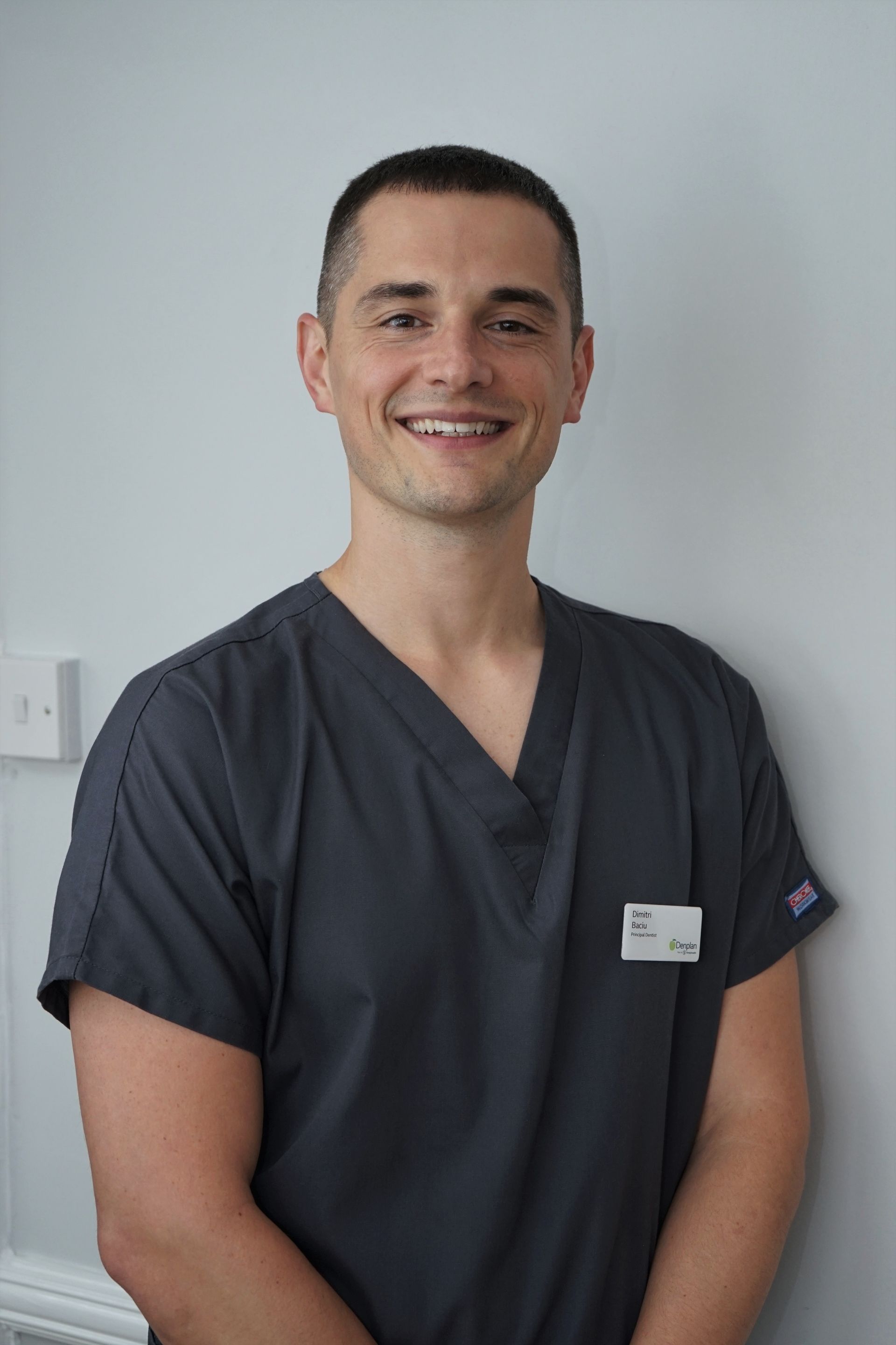 Man in grey scrubs smiles, standing against a white wall. He has short brown hair, light skin, and a name tag.