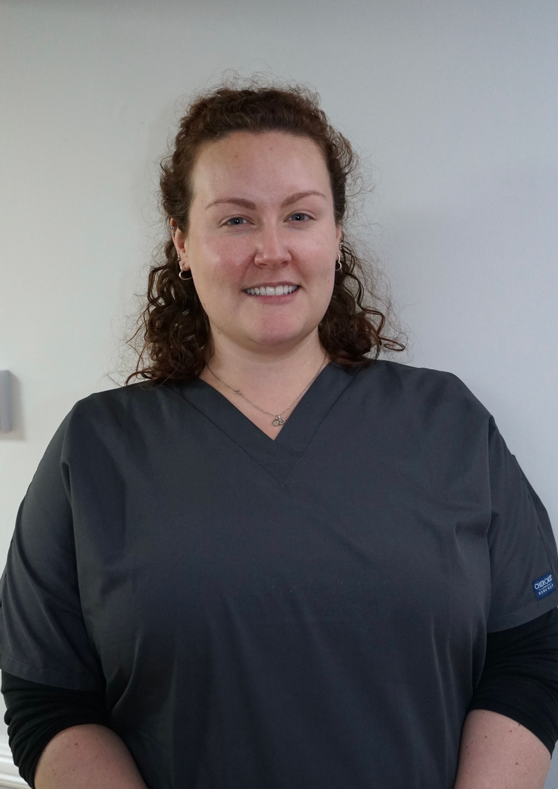 Woman with curly hair, wearing gray scrubs, smiling in a medical office setting.