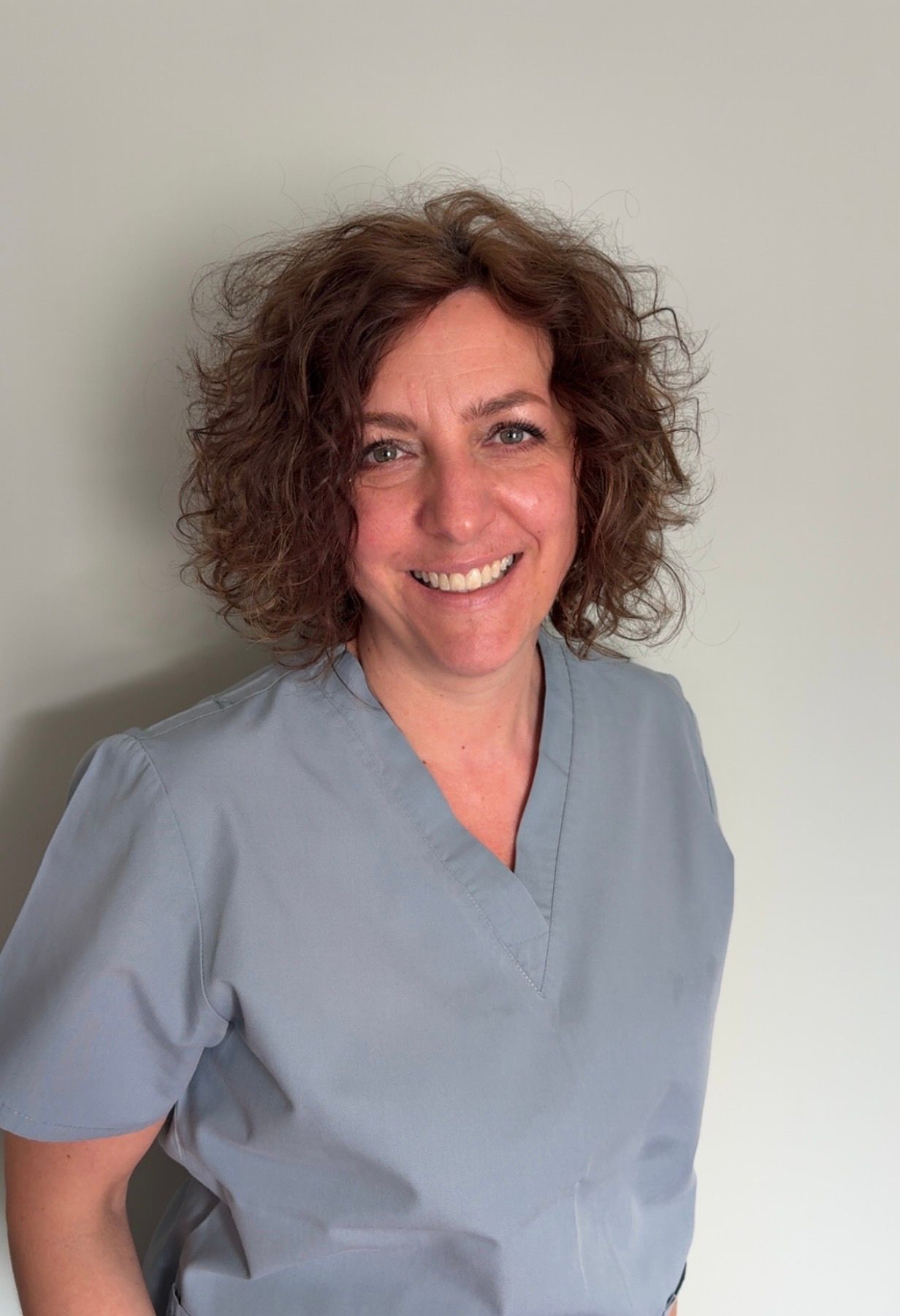 Woman in blue scrubs smiling; indoors against a white wall.