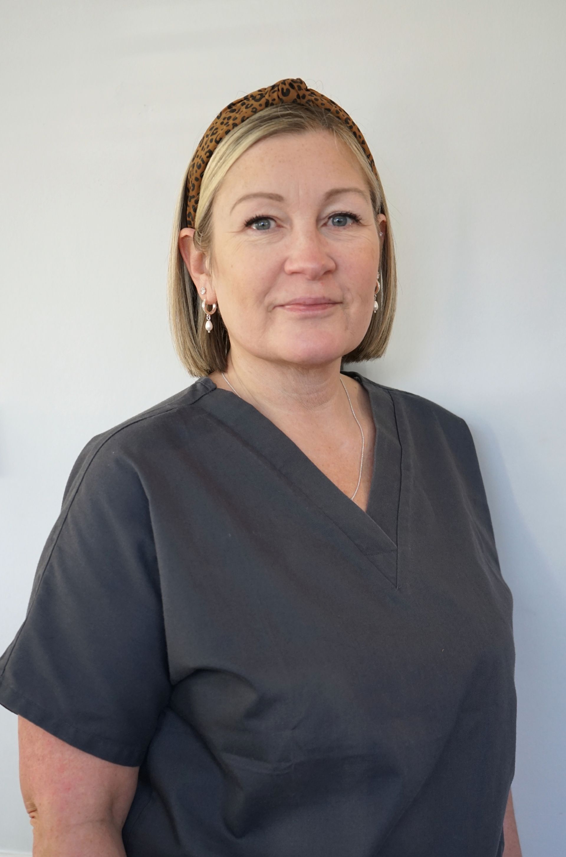 Woman in gray scrubs, wearing a headband, stands in front of a white wall.