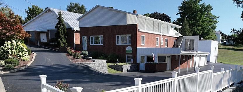 Brick apartment building with balconies beside a paved driveway and parked cars on a sunny day