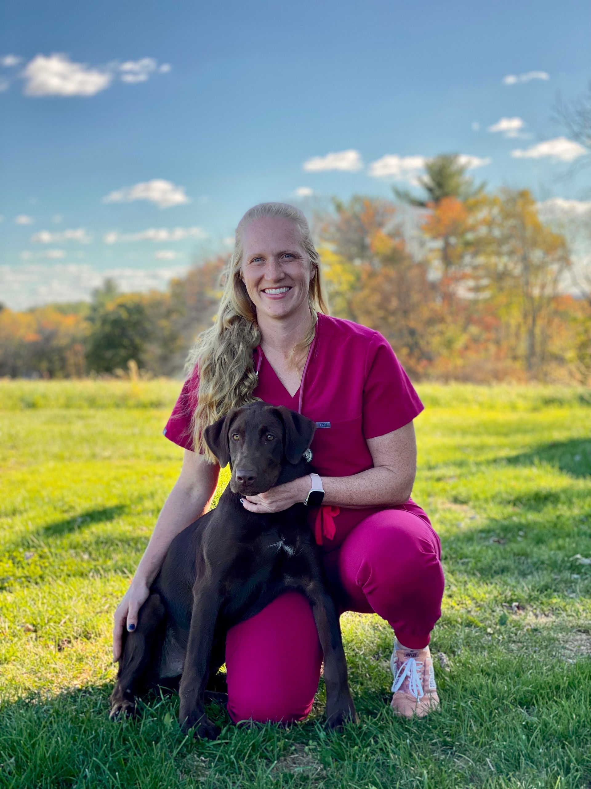 Dr. Hoffman in pink scrubs kneeling in a grassy field, smiling while holding a chocolate Labrador retriever.