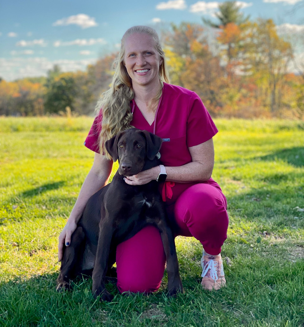 Dr. Hoffman in pink scrubs kneeling in a grassy field, smiling while holding a chocolate Labrador retriever.