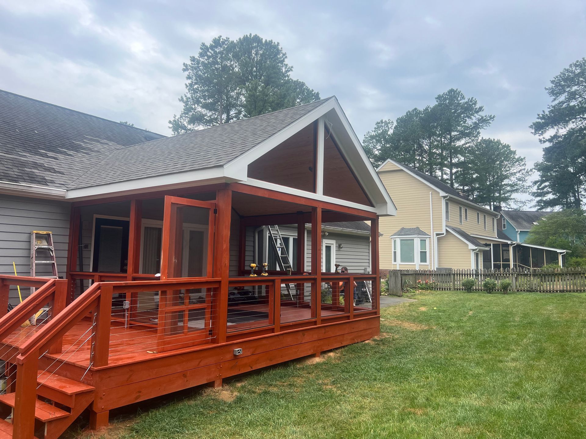 Red wooden deck and screened porch on a gray house, with a grassy yard and neighboring houses.