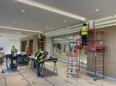 Construction workers install panels on a wall with a window. They use tools, lifts, and ladders in an unfinished space.