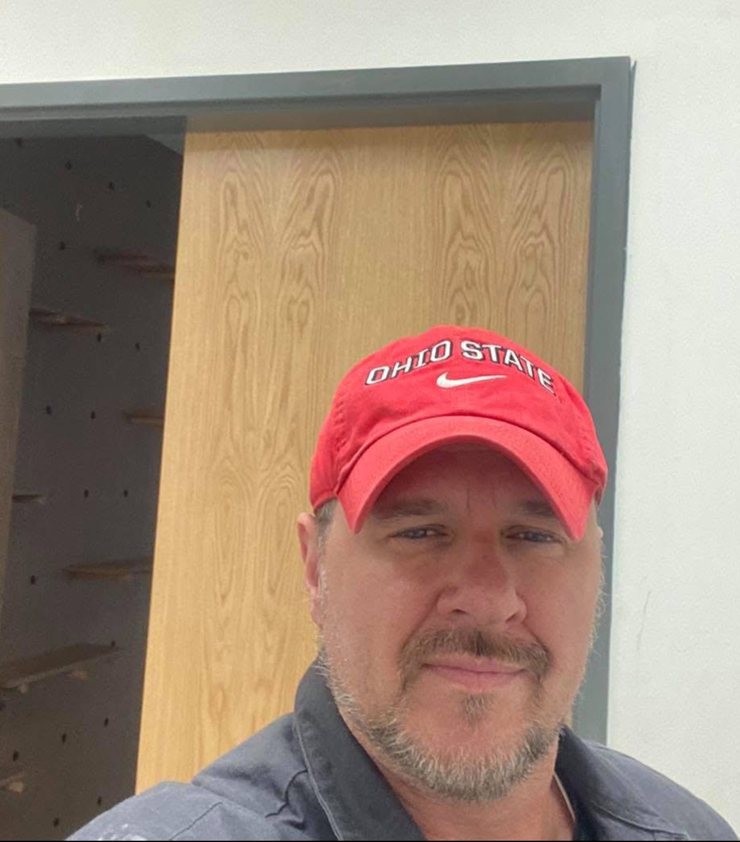 Man wearing an Ohio State hat, smiling, standing in front of a wooden door.
