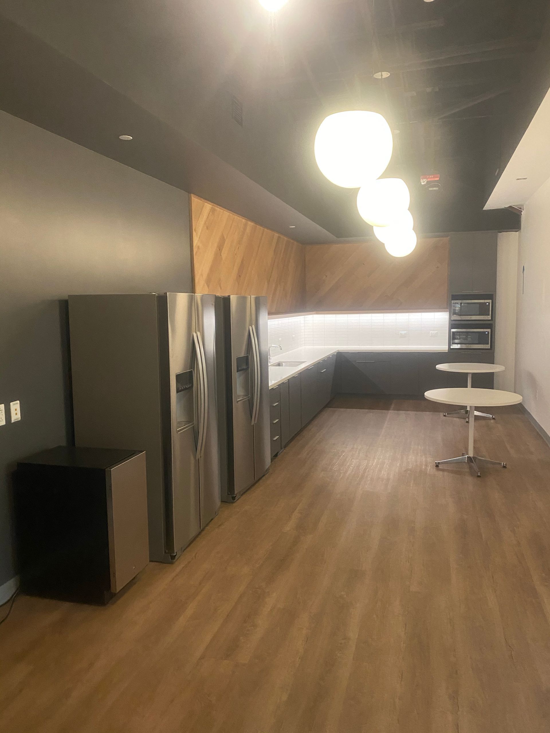 Modern office kitchen with fridges, cabinets, and round tables under globe lights.
