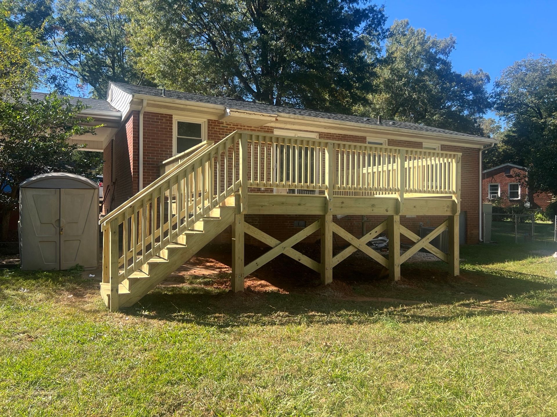 A wooden deck with stairs attached to a brick building, supported by X-shaped braces, on green grass.