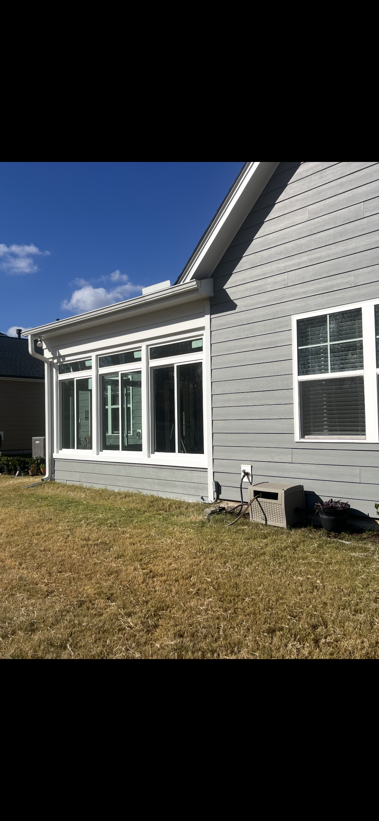 Exterior view of a house with gray siding, white windows, and a sunroom with a grassy lawn under a blue sky.