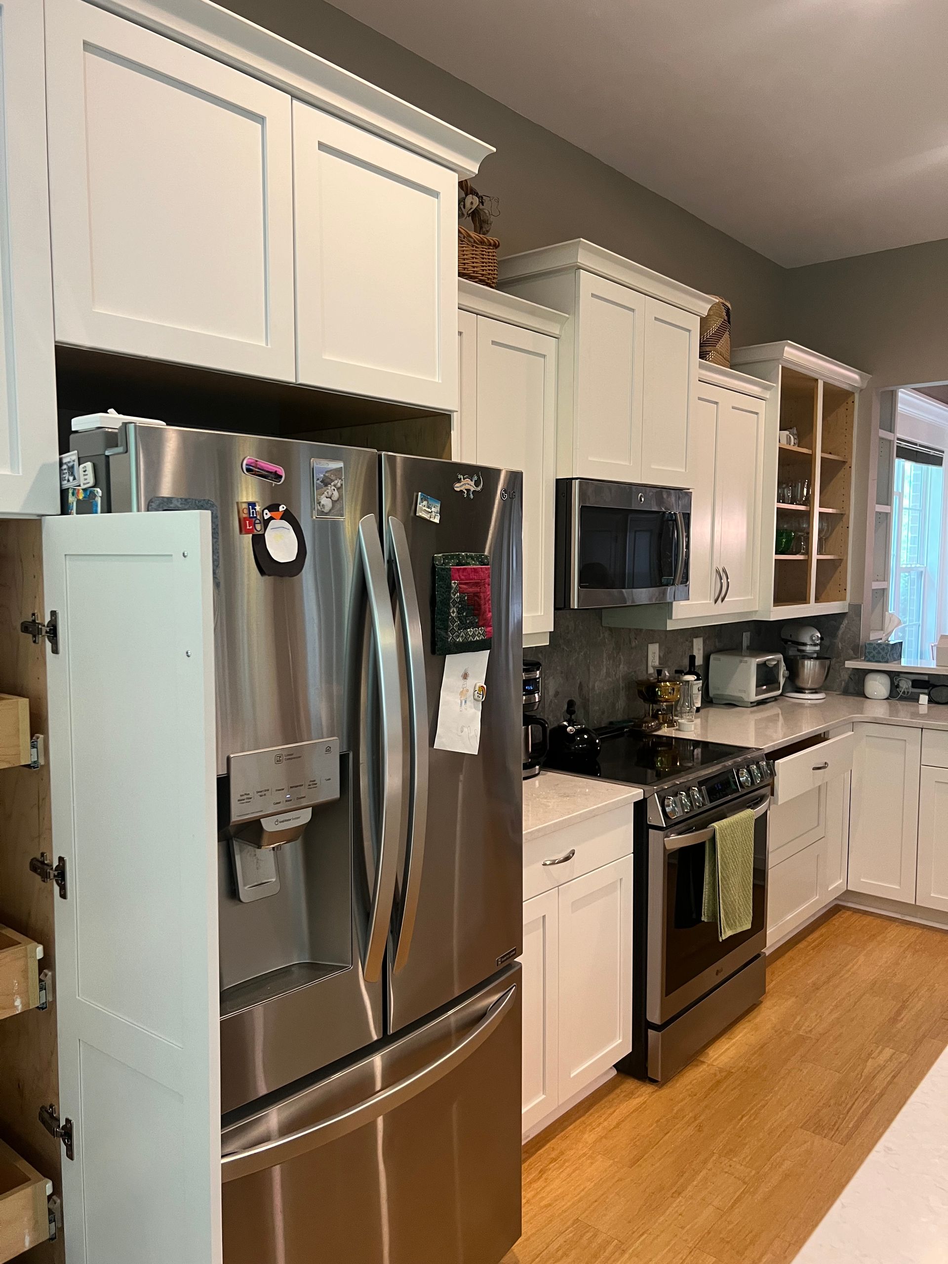 White kitchen cabinets frame a stainless steel refrigerator and appliances. Light wood floors and counters.