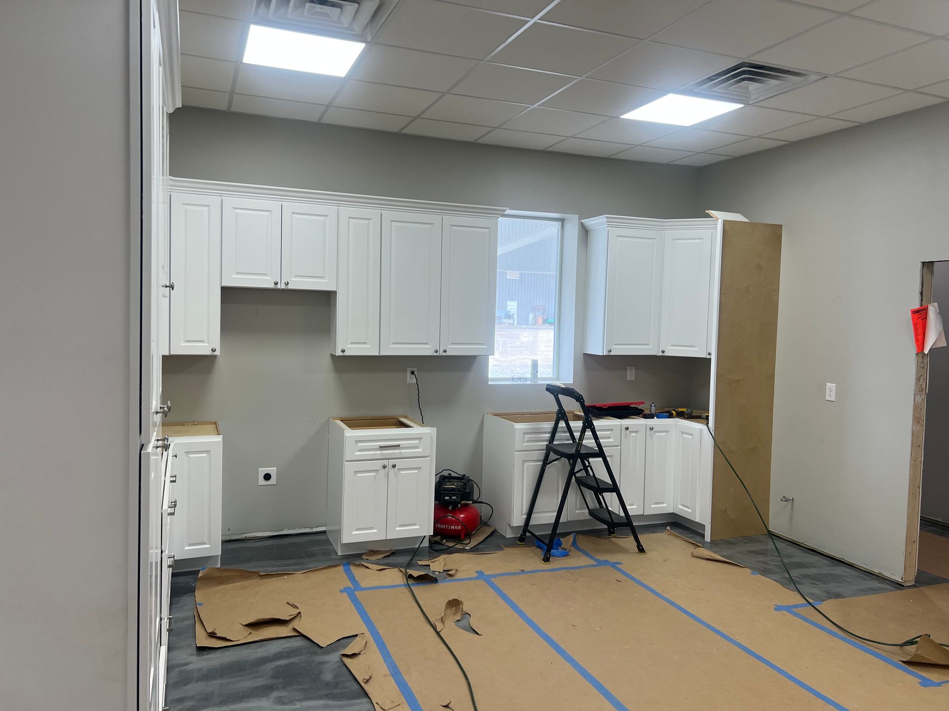 Kitchen cabinets being installed in a room. White cabinets, grey walls, cardboard on floor.