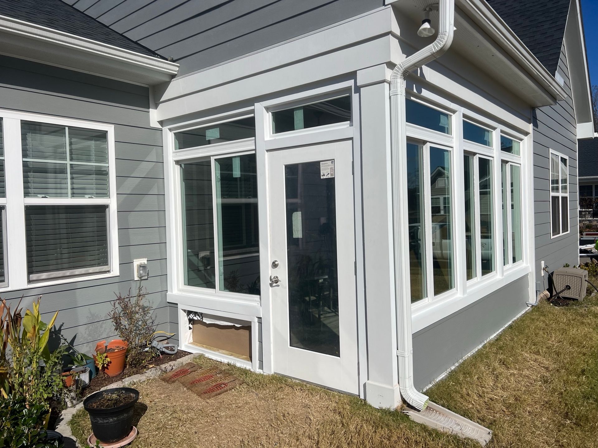 Exterior view of a gray house with a white-framed sunroom and door, surrounded by grass and plants.