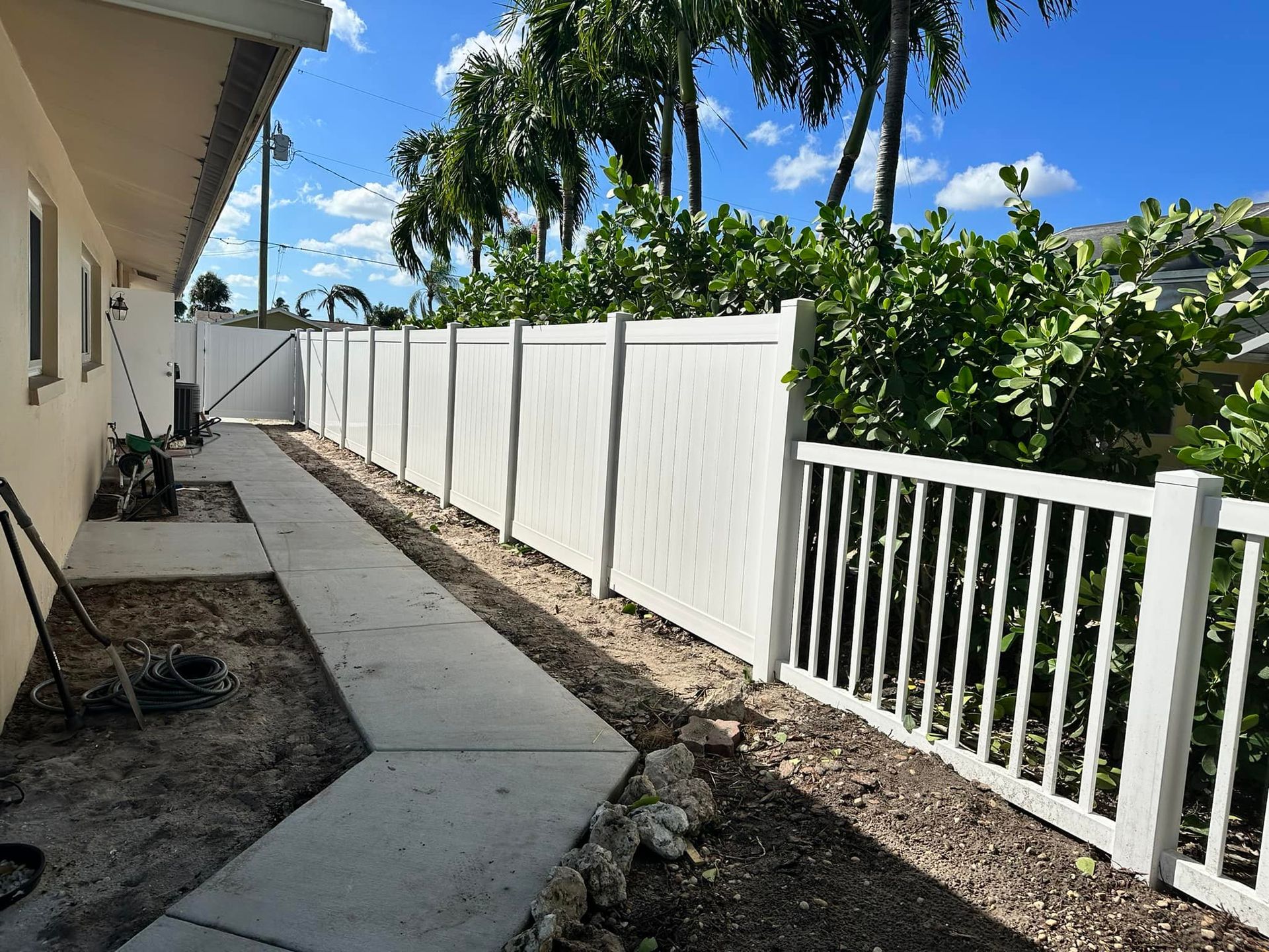 White fence alongside a concrete walkway next to a building, with lush greenery and blue sky in the background.
