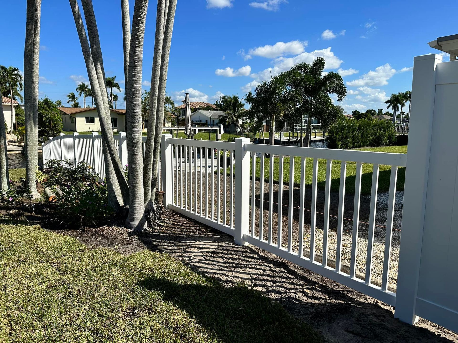 White fence bordering green lawn with palm trees, blue sky, and distant houses.