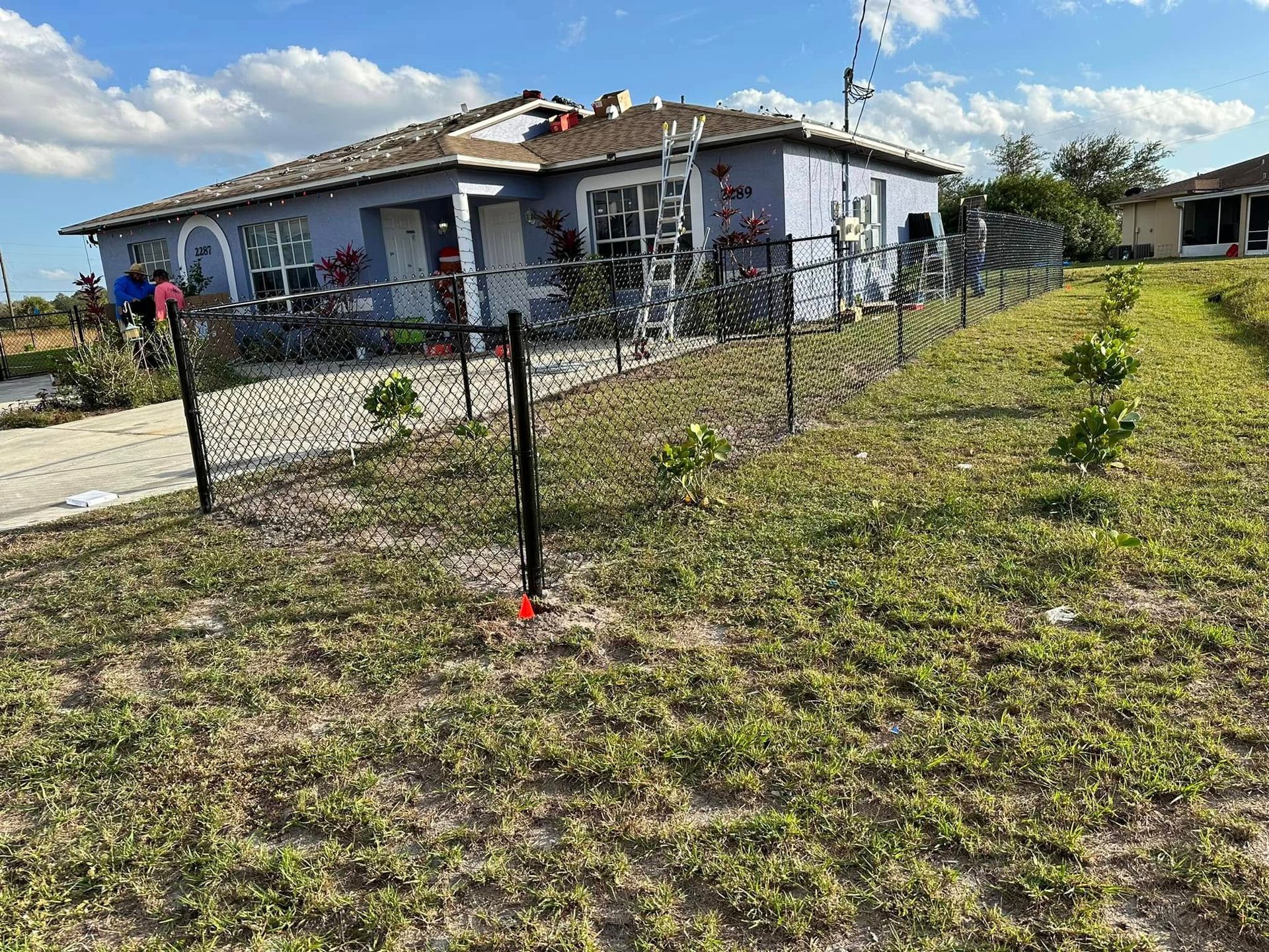 A light blue house with a black chain link fence in front. Green grass and blue sky.
