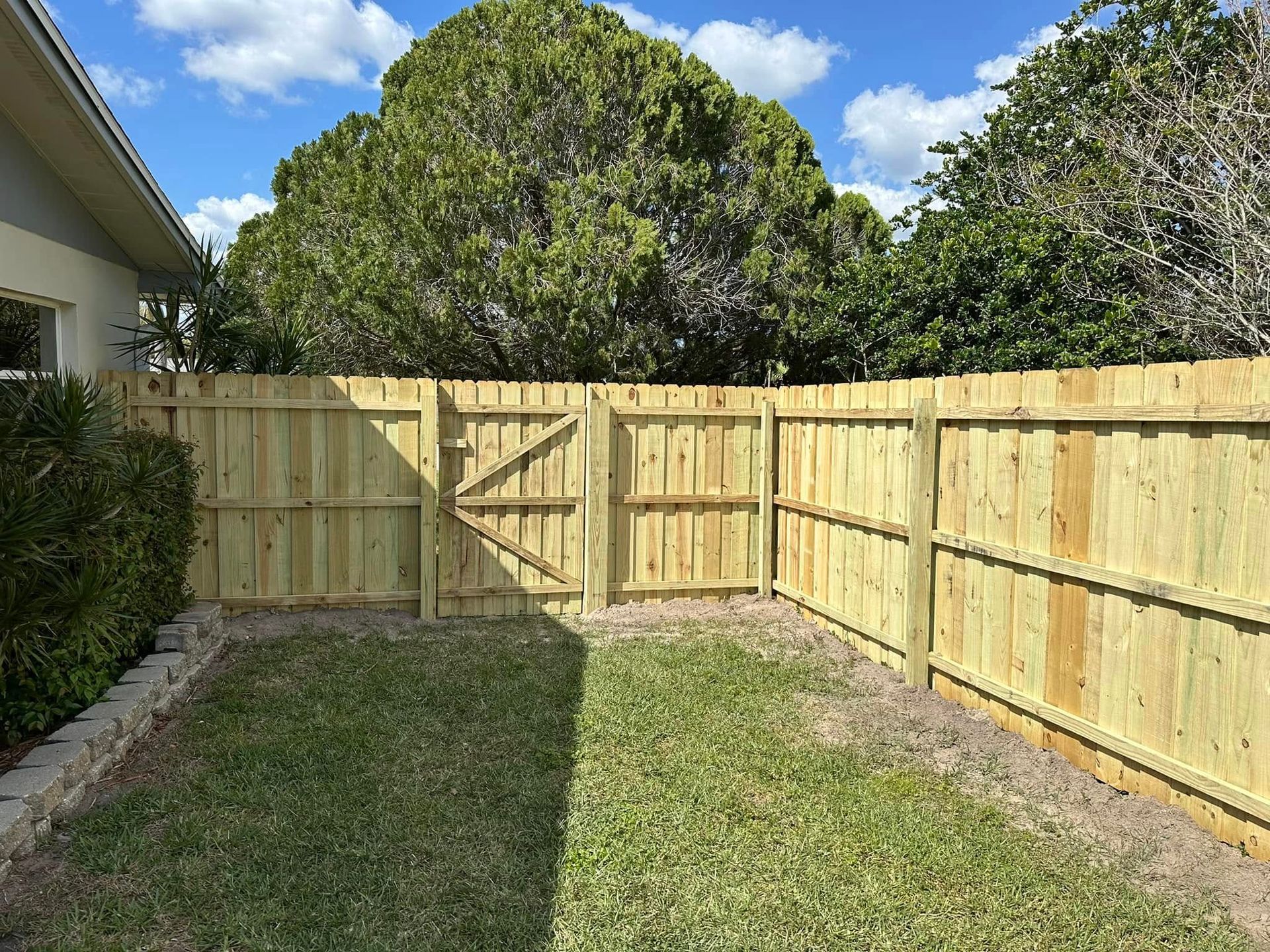 Wooden fence with gate in a yard with green grass, trees, and a building.