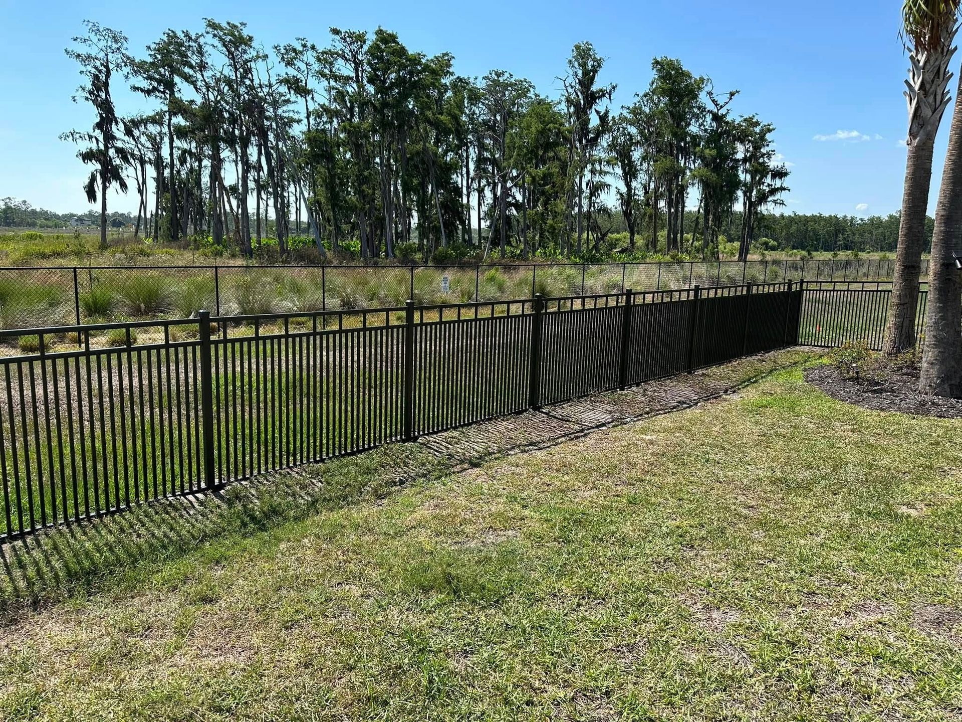 Black fence along a grassy slope, with trees and blue sky in the background.