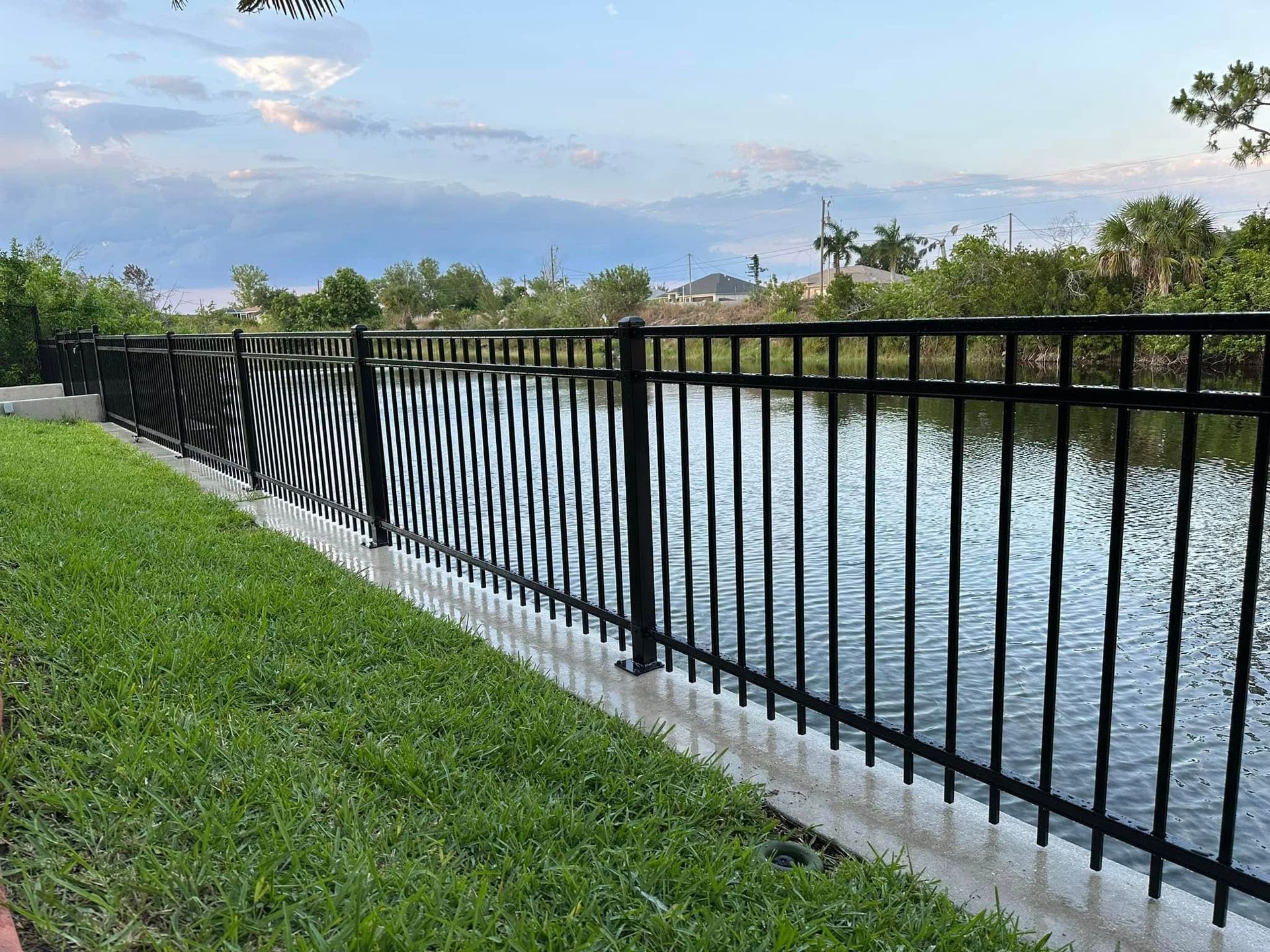 Black metal fence along a waterway, with green grass in the foreground and cloudy sky above.