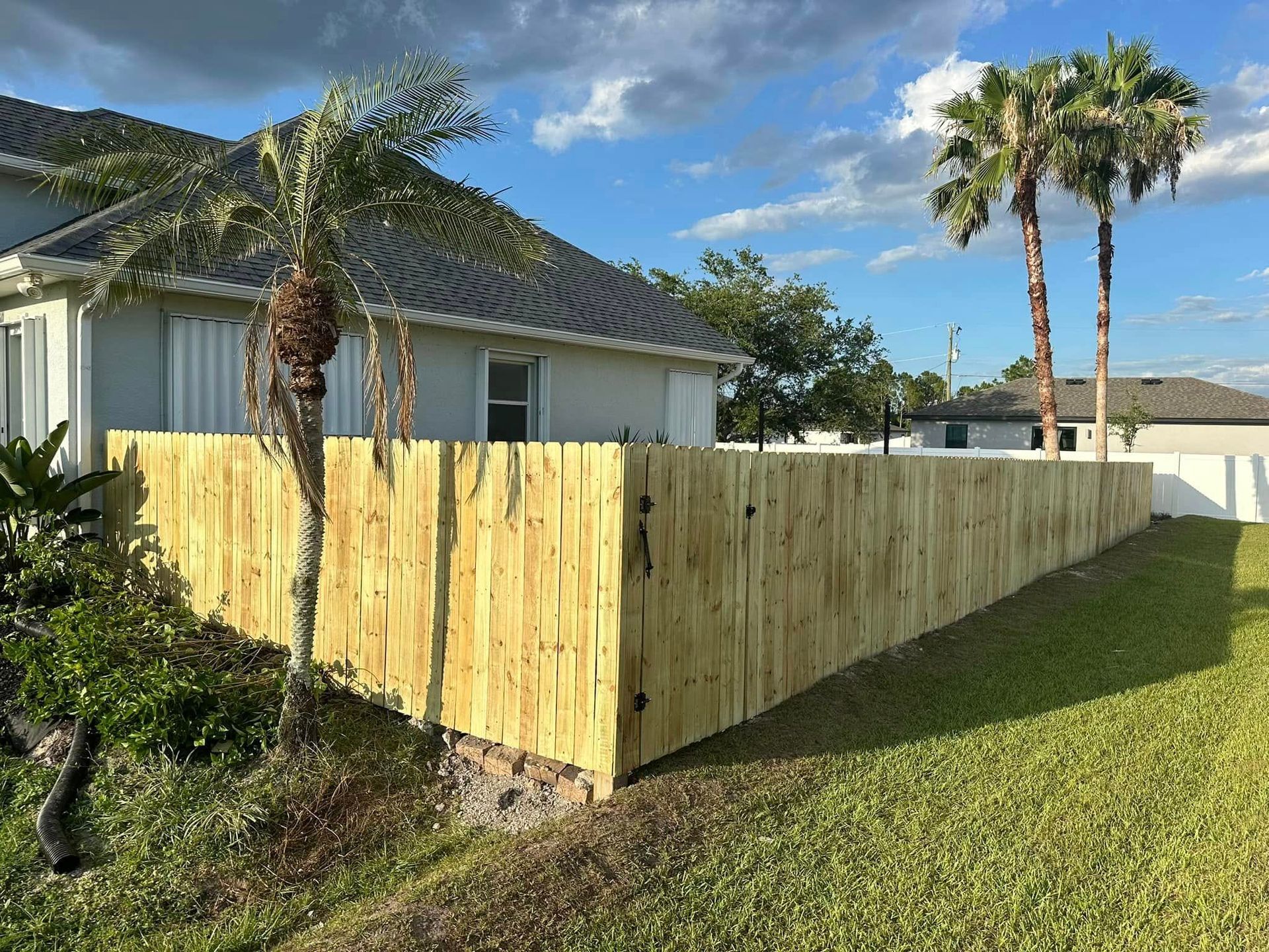 Wooden fence along a grassy yard, next to a light-colored house with palm trees under a partly cloudy sky.