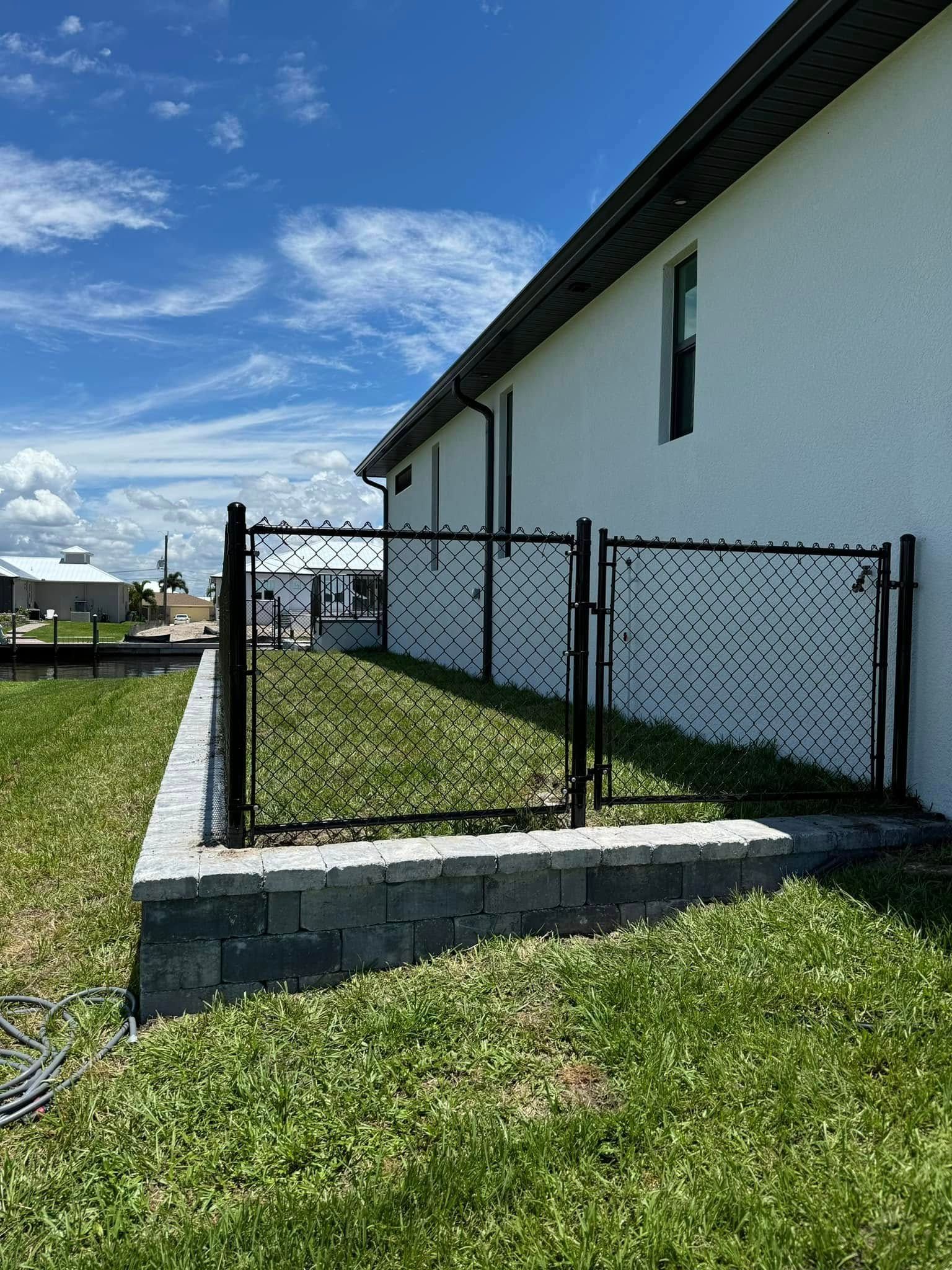Black metal fence on a gray block retaining wall. Green grass, white house, and blue sky.