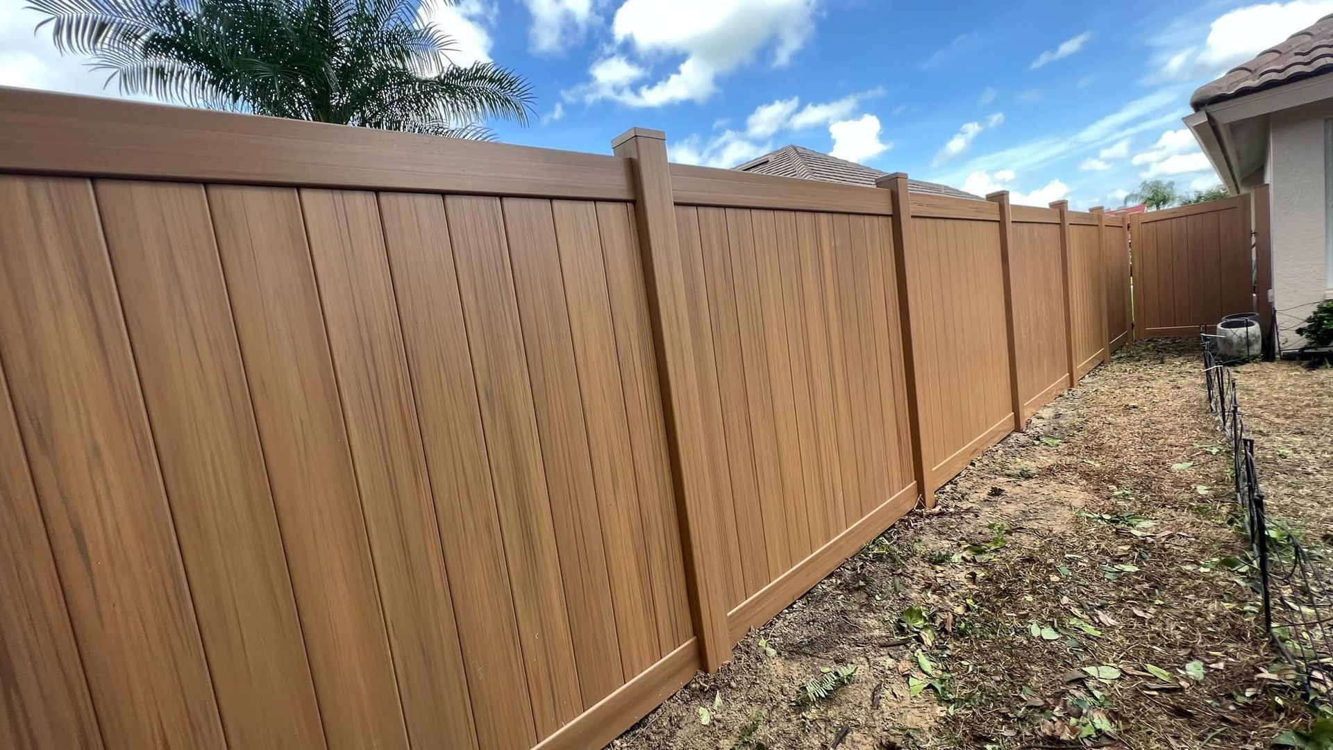 Brown vinyl fence bordering a yard under a partly cloudy blue sky.