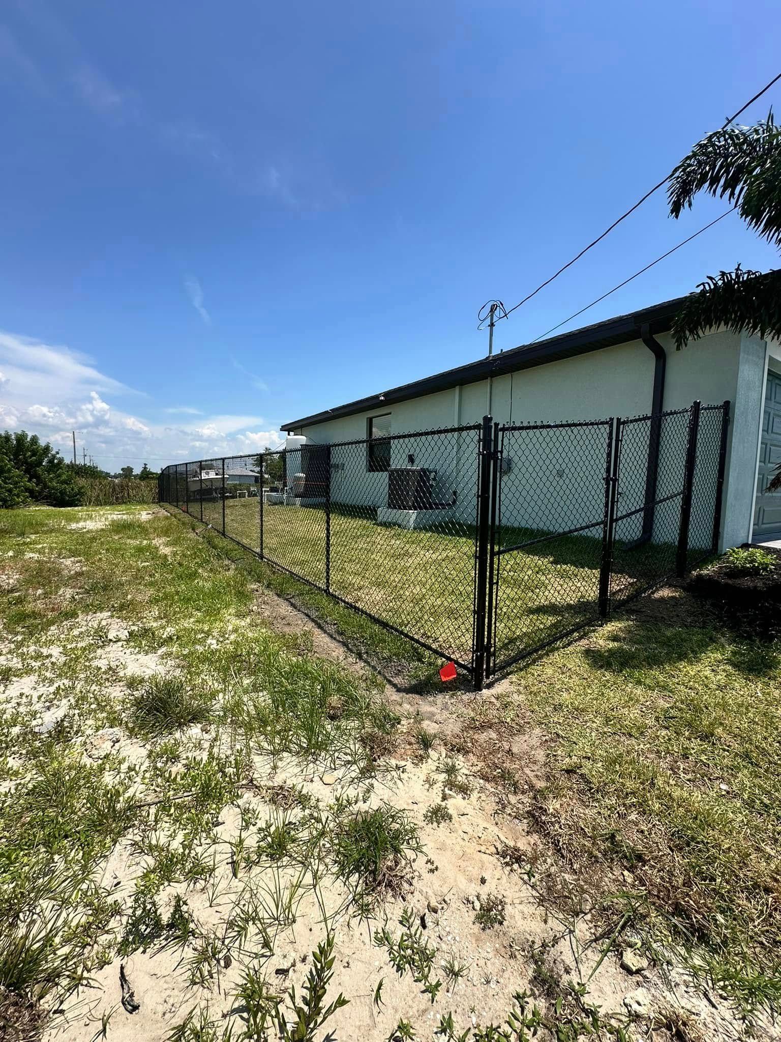 A black metal fence encloses a white building on a sunny day; overgrown grass in the foreground.