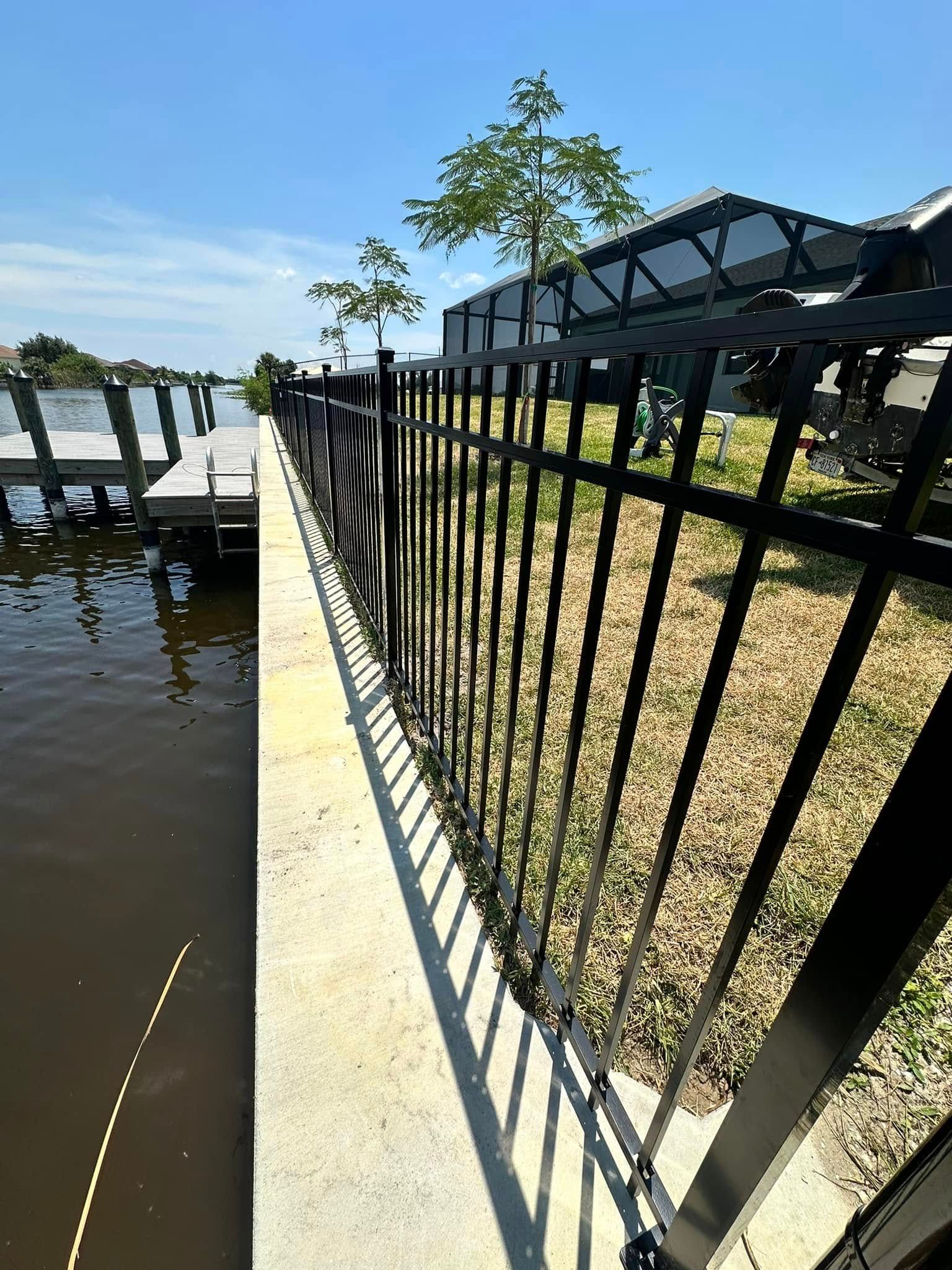 Black metal fence along a concrete walkway next to a waterway with a dock on a sunny day.